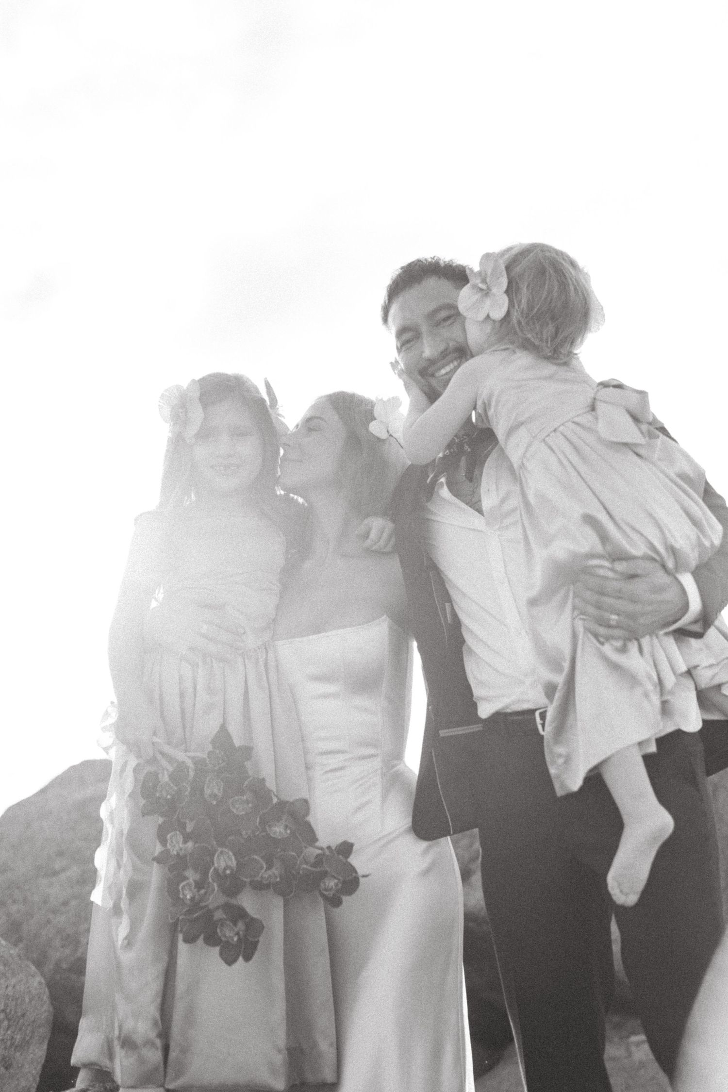 Wedding party guests in formal attire dance and embrace outdoors with bright sunlight behind them.