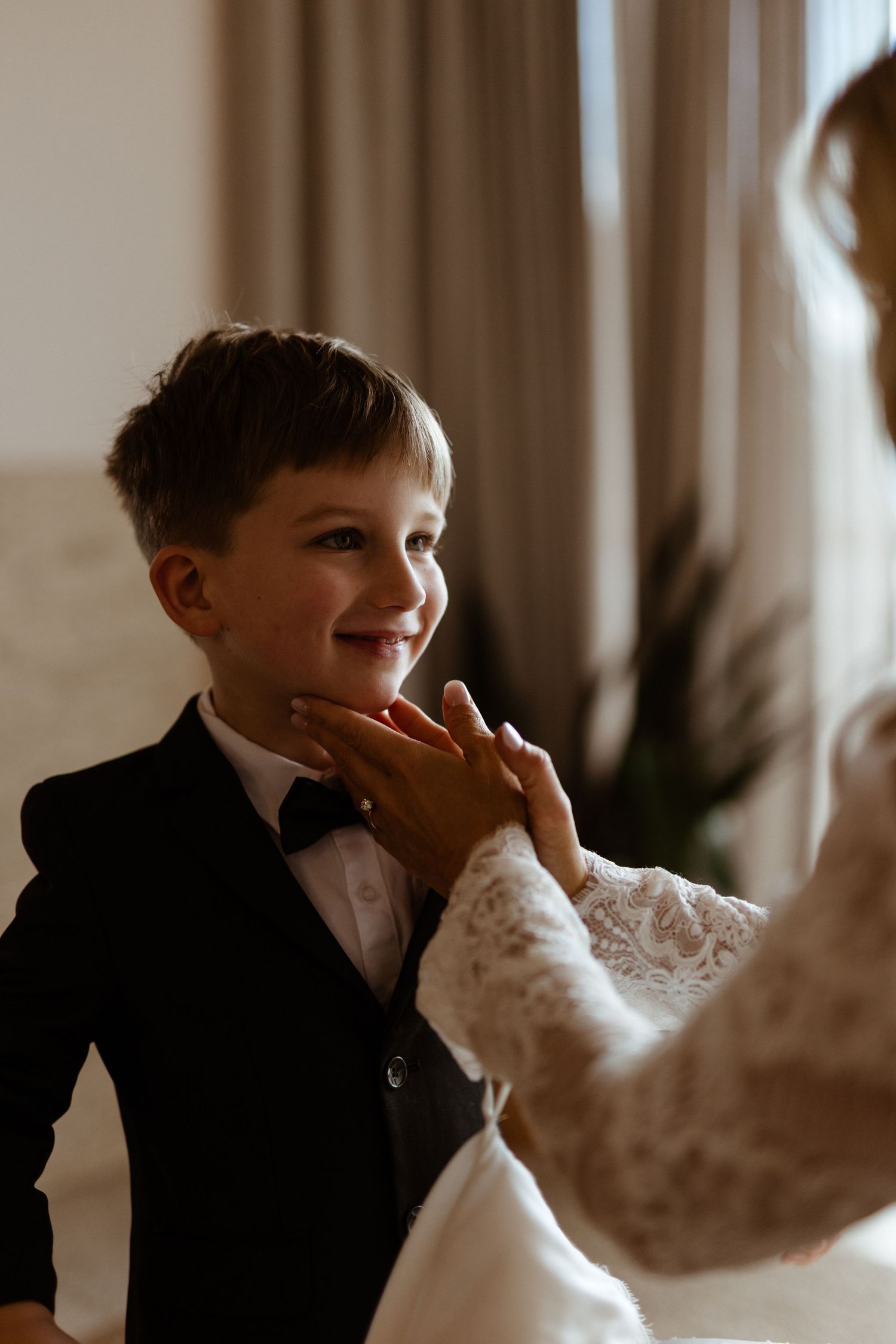 A young boy in a black tuxedo smiles while someone in a lace sleeve gently touches his face.