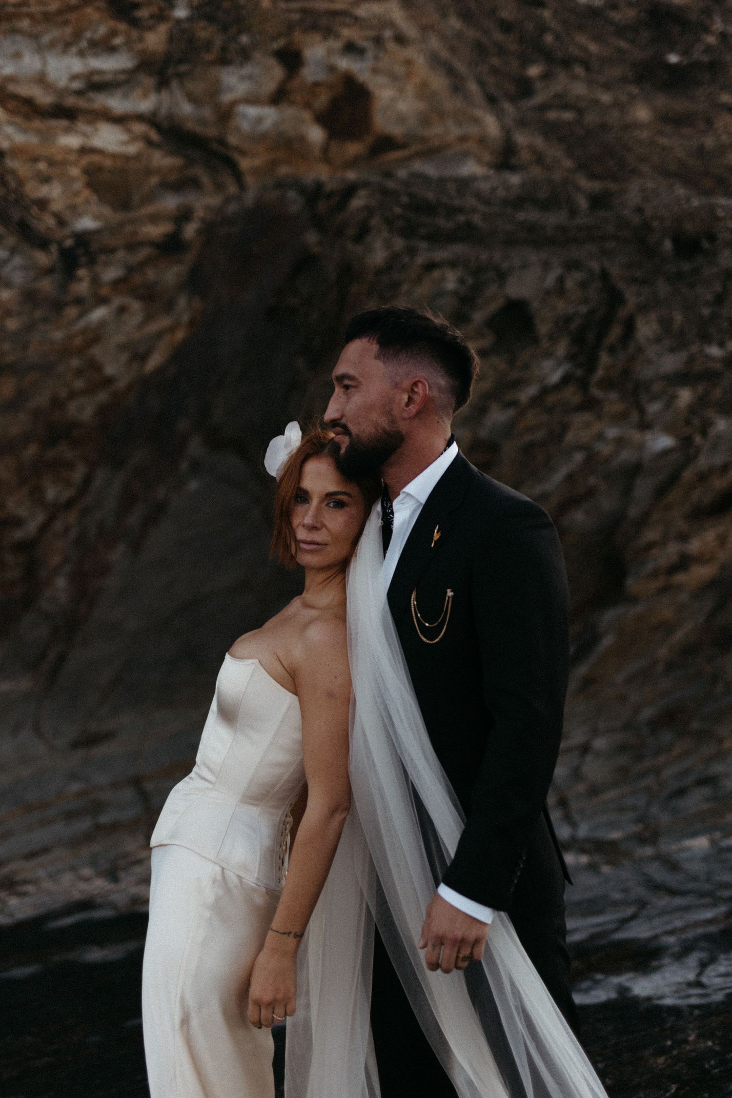 A bride and groom pose together against dramatic rocky cliffs at sunset in elegant wedding attire.