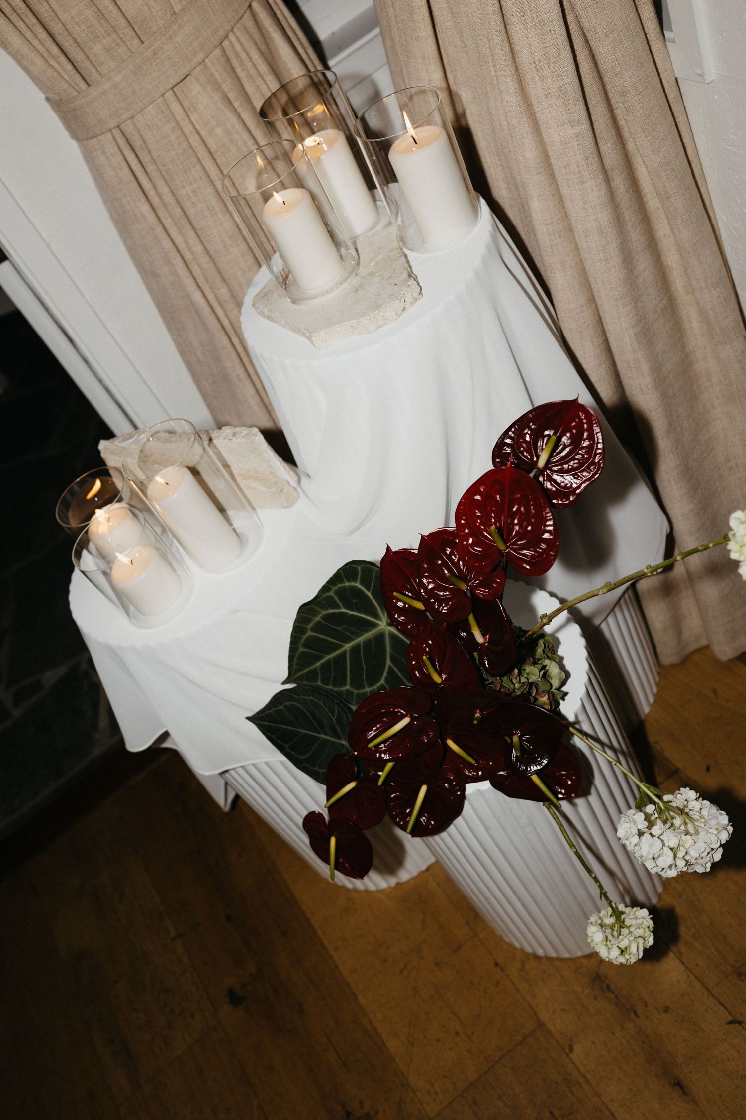 Dark burgundy anthurium flowers with waxy petals rest in a white ceramic vase against a wooden floor and curtained wall.