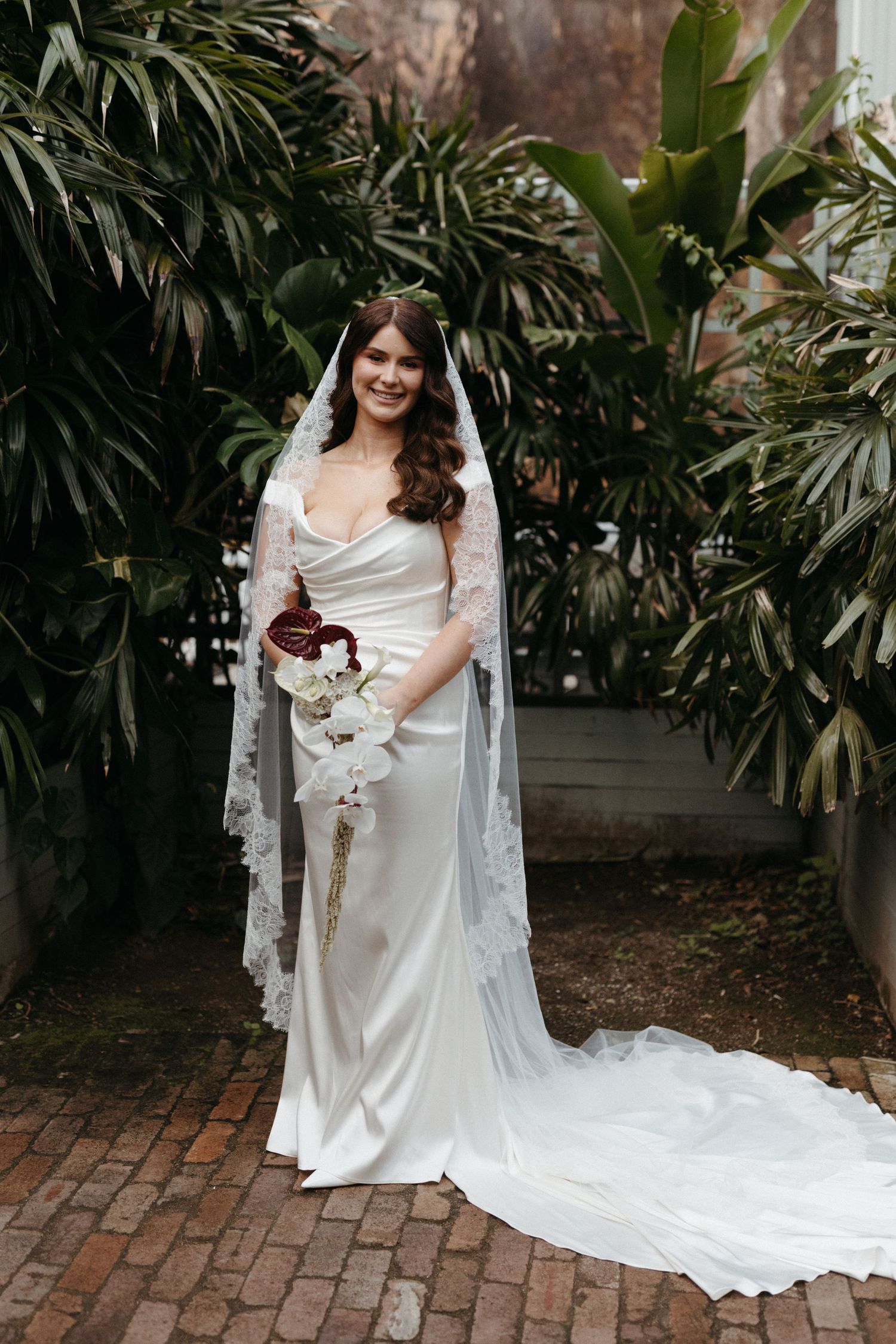 A bride in flowing white gown and lace veil poses against lush tropical greenery and brick path.