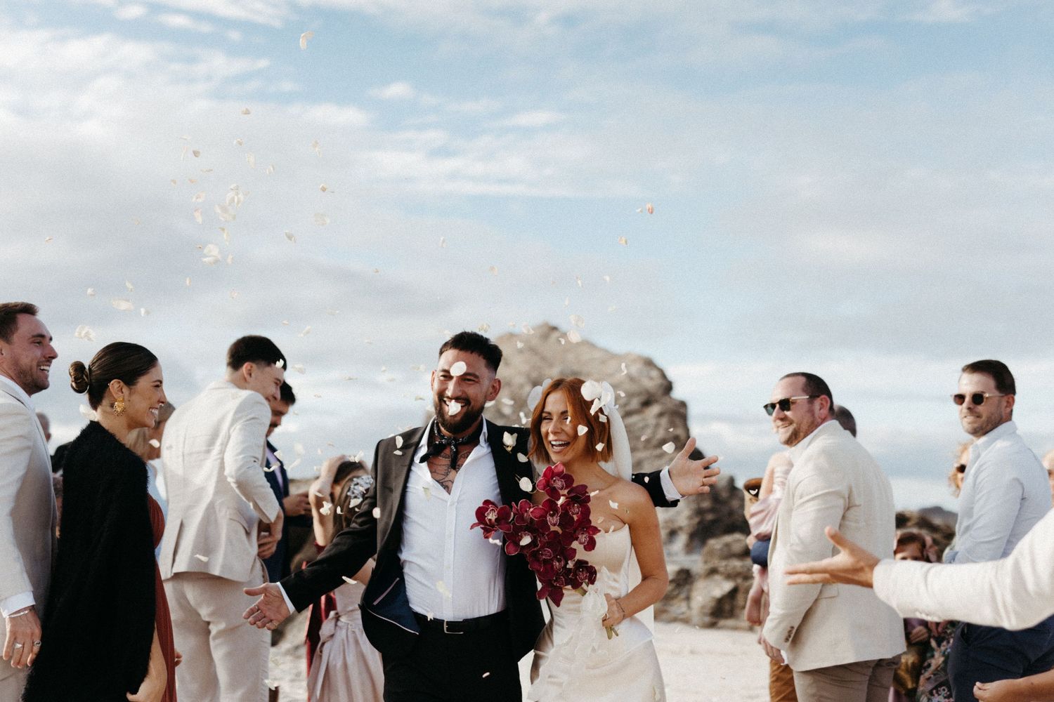 Newlywed couple celebrates with wedding party on a scenic cliffside beach under cloudy skies.