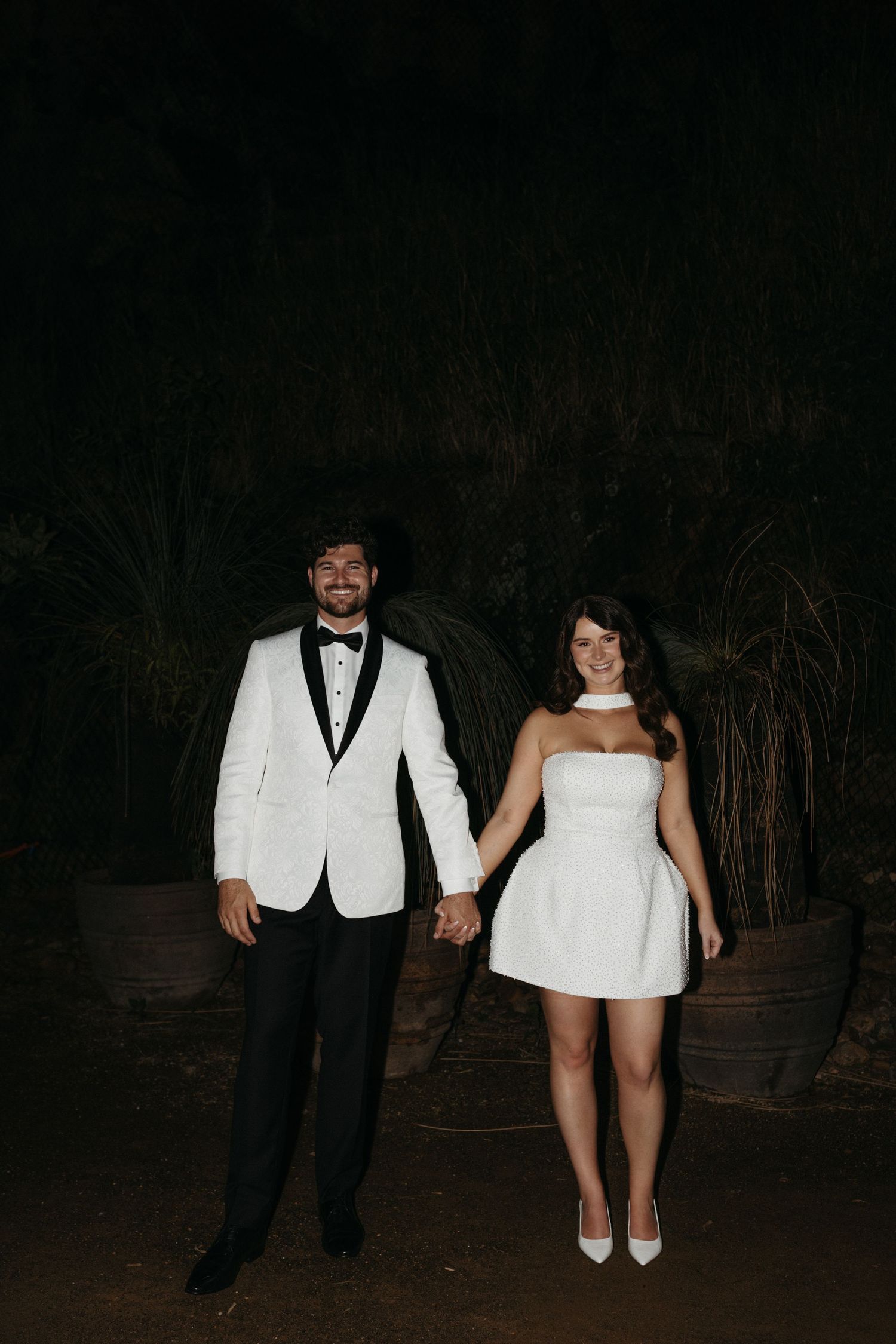 A couple in formal wedding attire pose together at nighttime in front of a dark wooded area.