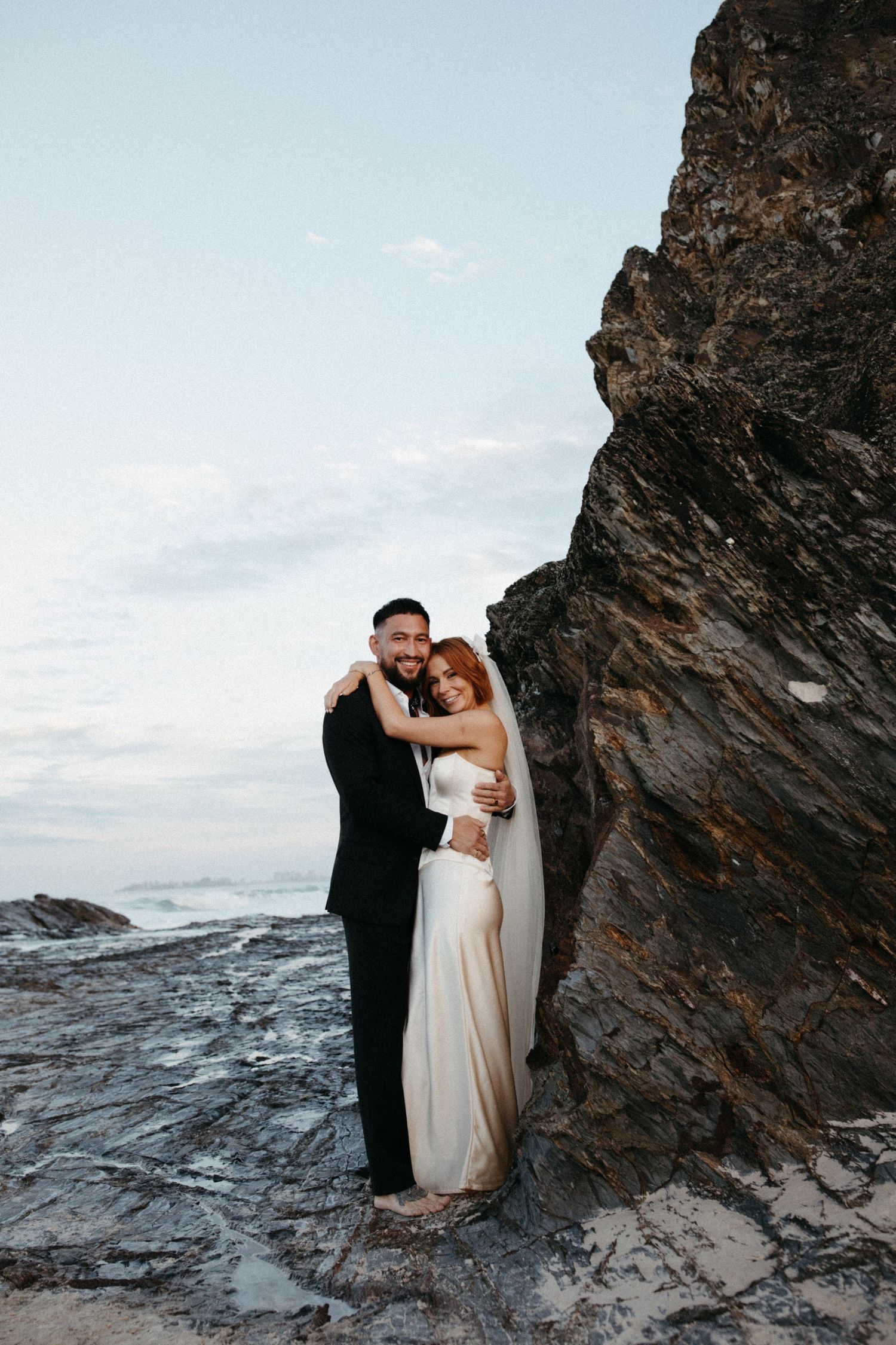A romantic couple embraces on rocky coastal cliffs during a seaside photo session at sunset.
