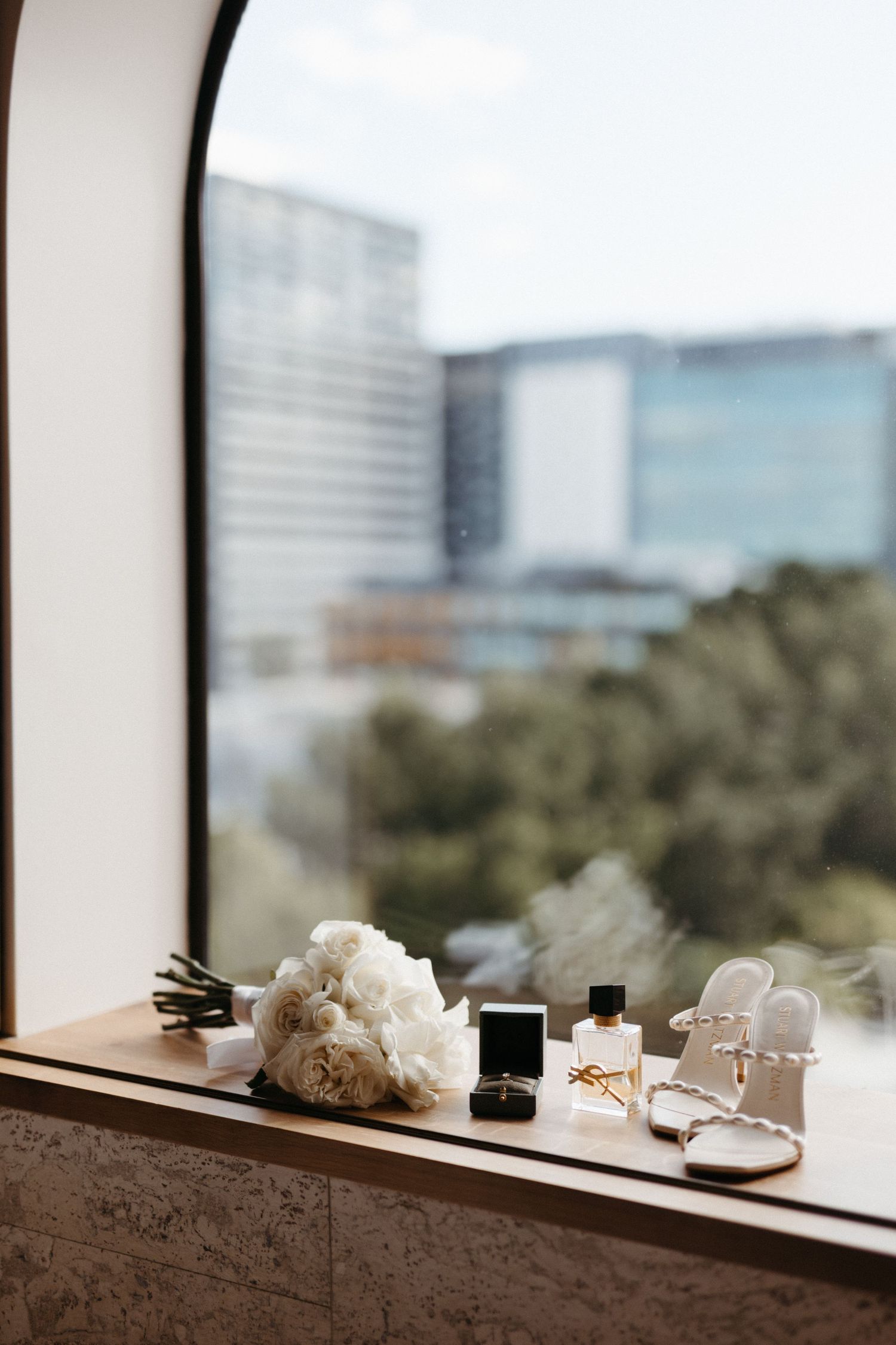 Wedding details on a window ledge include white roses, perfume, rings and jewelry with blurred cityscape in background.