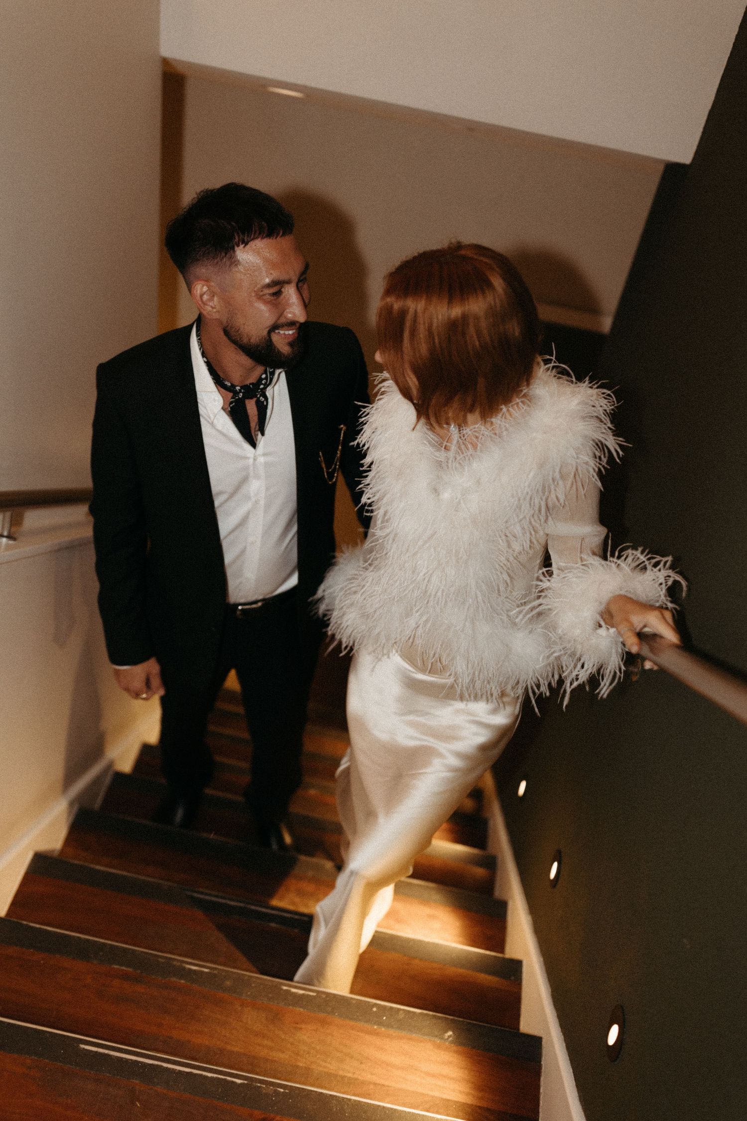 Couple in formal attire descend wooden staircase, bride wearing feathered white dress.