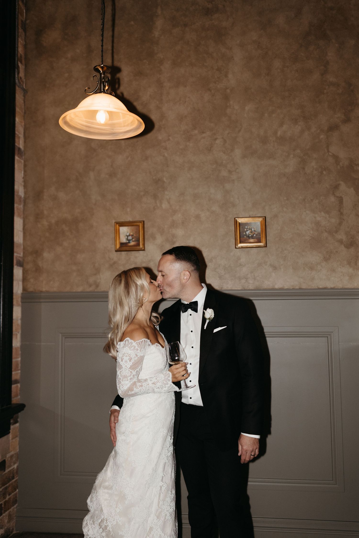 A couple shares an intimate moment in formalwear under a vintage pendant light against textured walls with framed artwork.