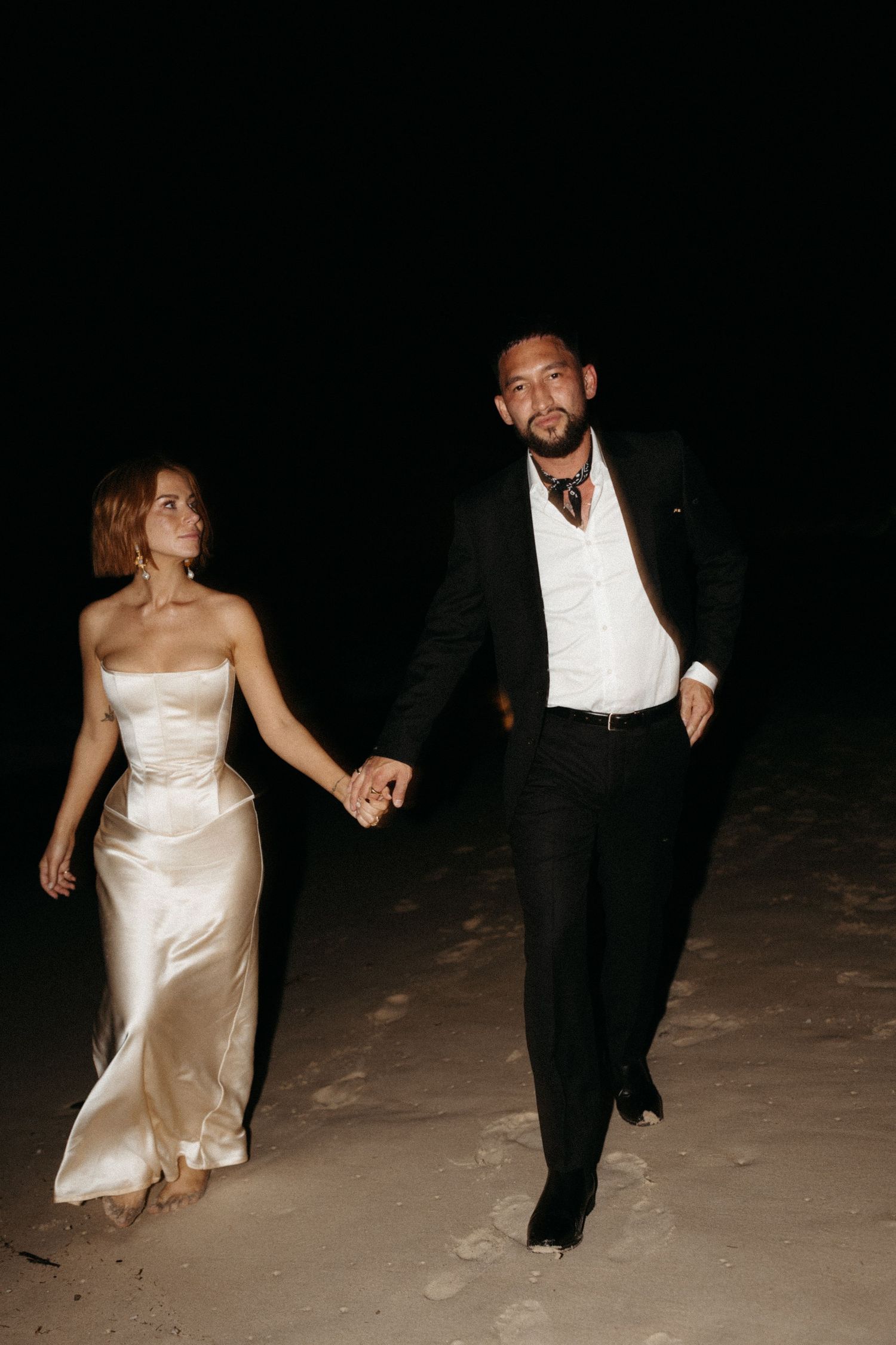 Newlyweds holding hands while walking on beach at night.