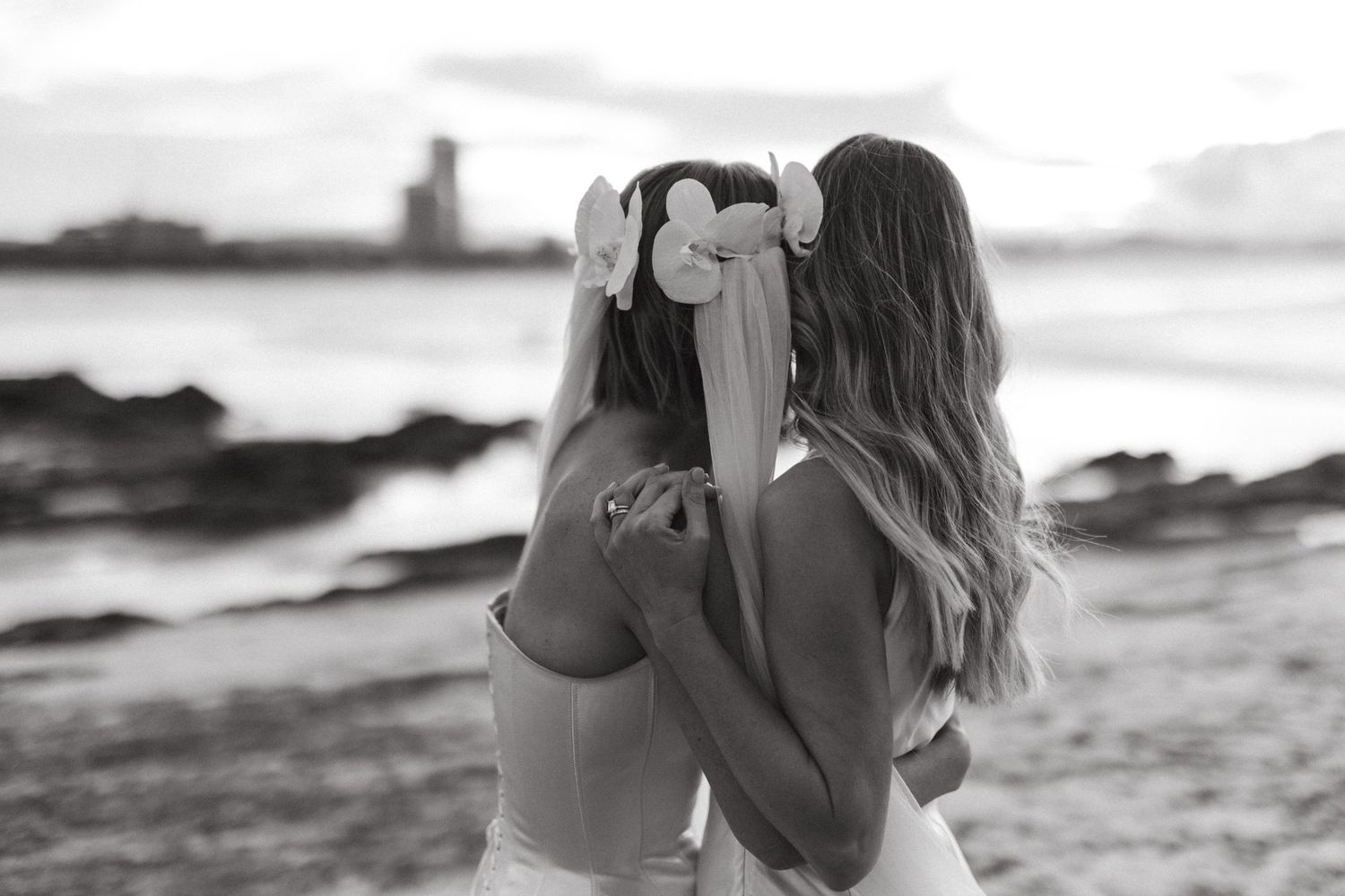 Two silhouettes embrace while standing on a rocky beach at sunset in a black and white photograph.