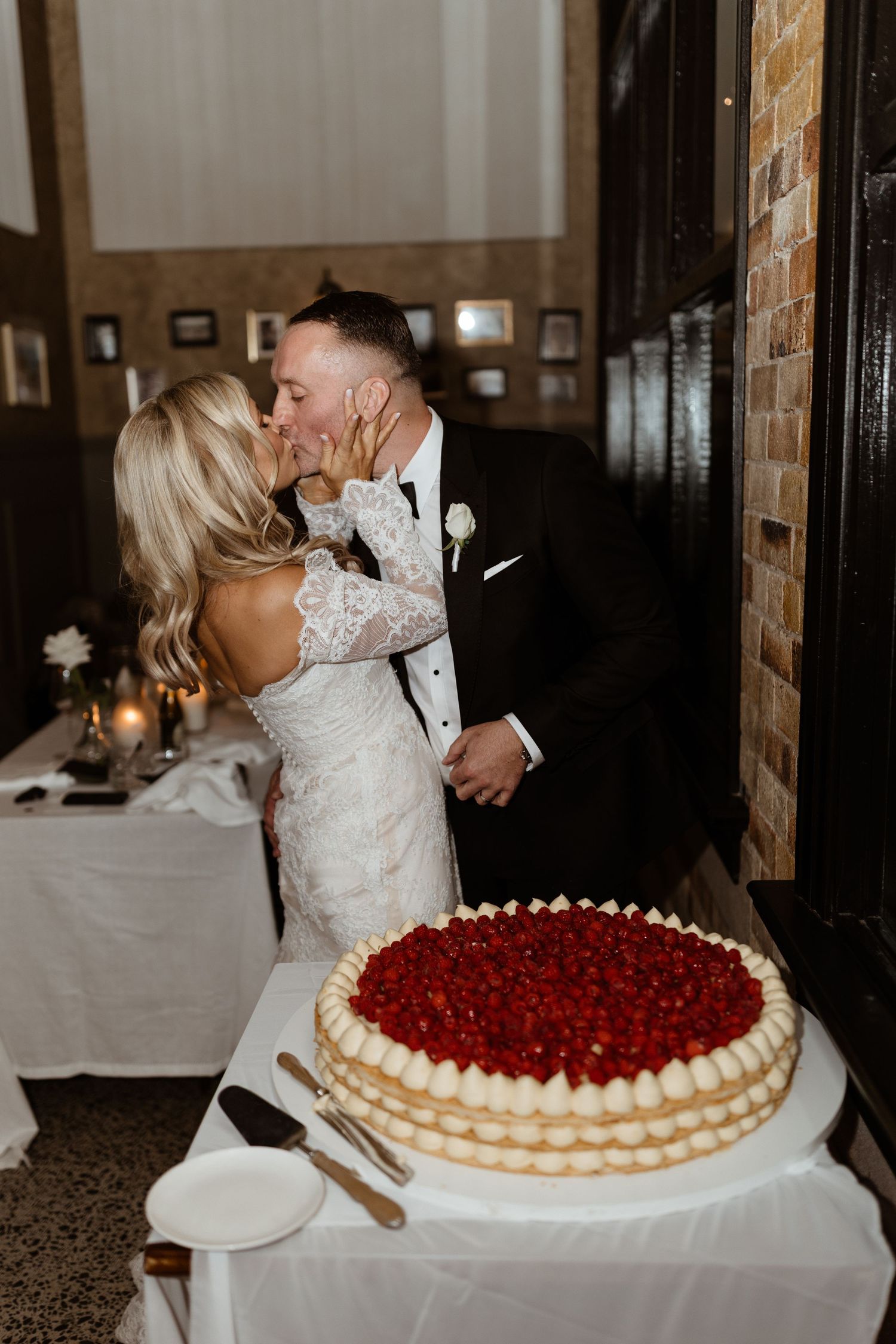 A wedding couple sharing a kiss beside a large layered cake topped with fresh red berries at their reception.