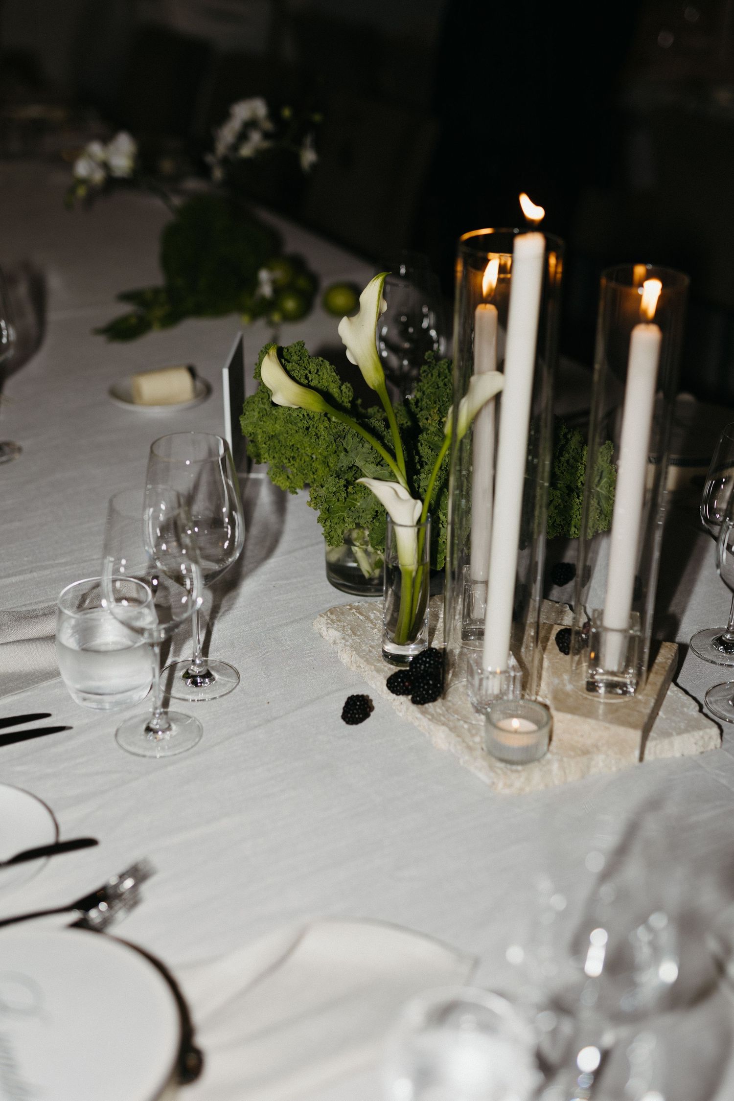 Elegant dinner table setting with white taper candles, calla lilies, and moss centerpiece on crisp white tablecloth.