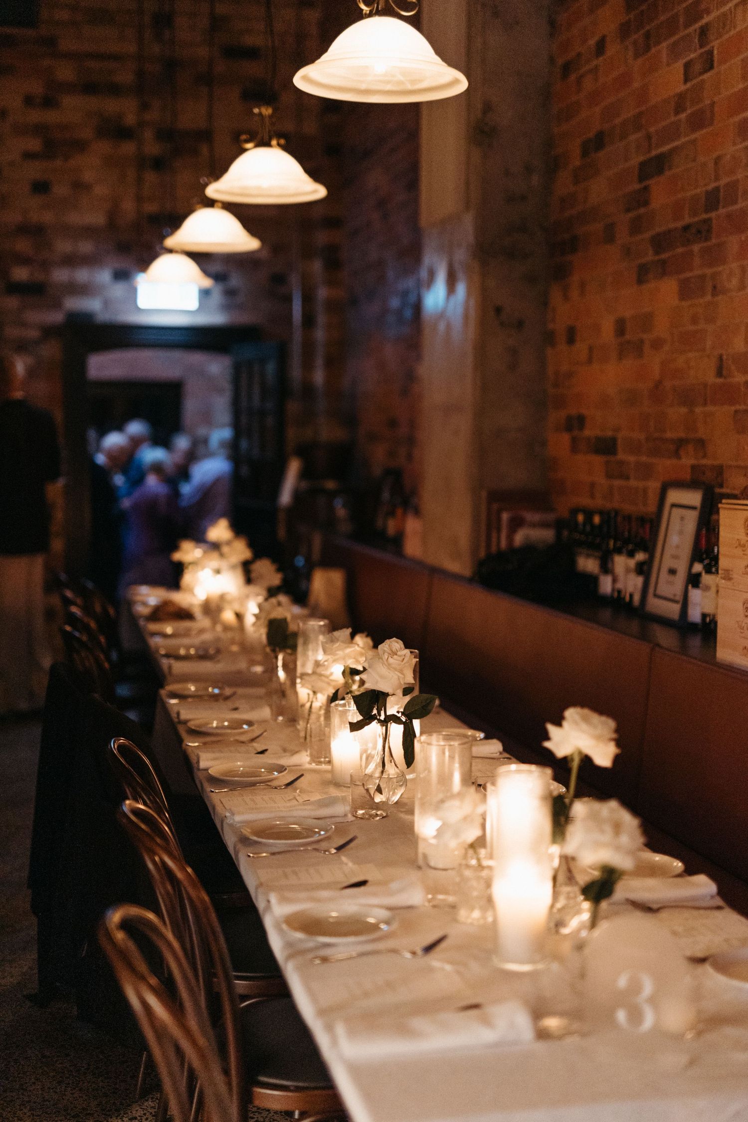 A long white-clothed dining table with elegant place settings and candlelight glows warmly against exposed brick walls.