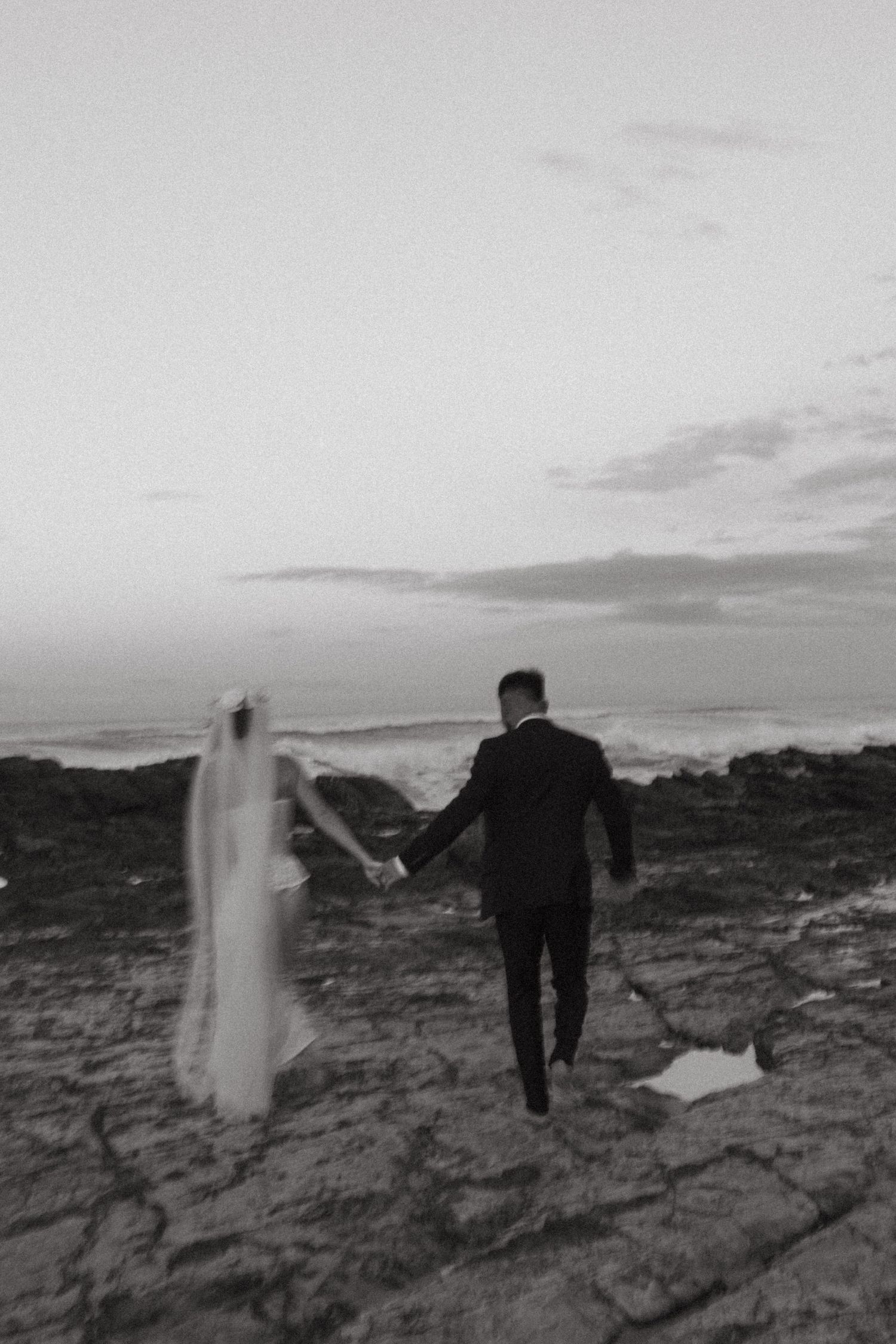 Black and white photo of couple walking hand in hand on rocky coastal landscape at sunset.