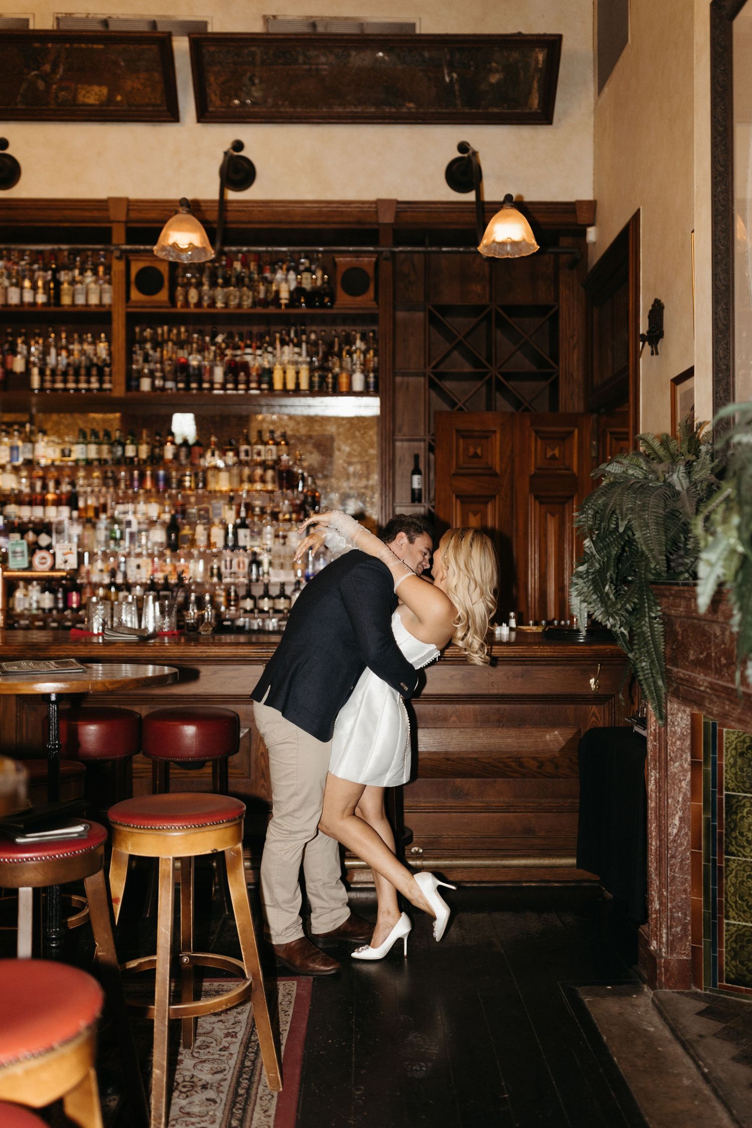 A romantic embrace in an intimate vintage bar setting with warm lighting and a well-stocked wooden back bar.
