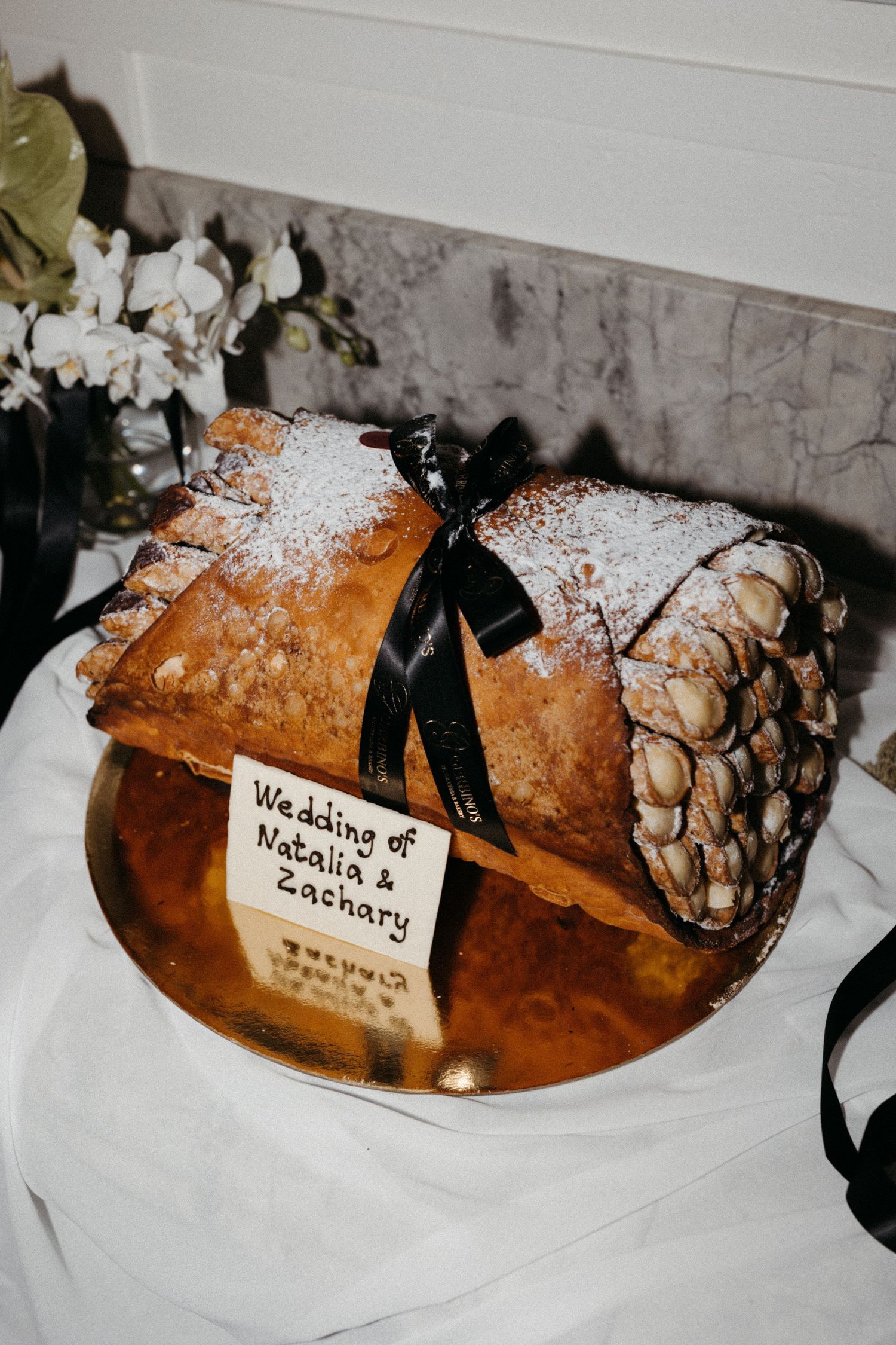 Wedding dessert with powdered sugar dusted pastries tied with black ribbons on a platter next to white flowers.