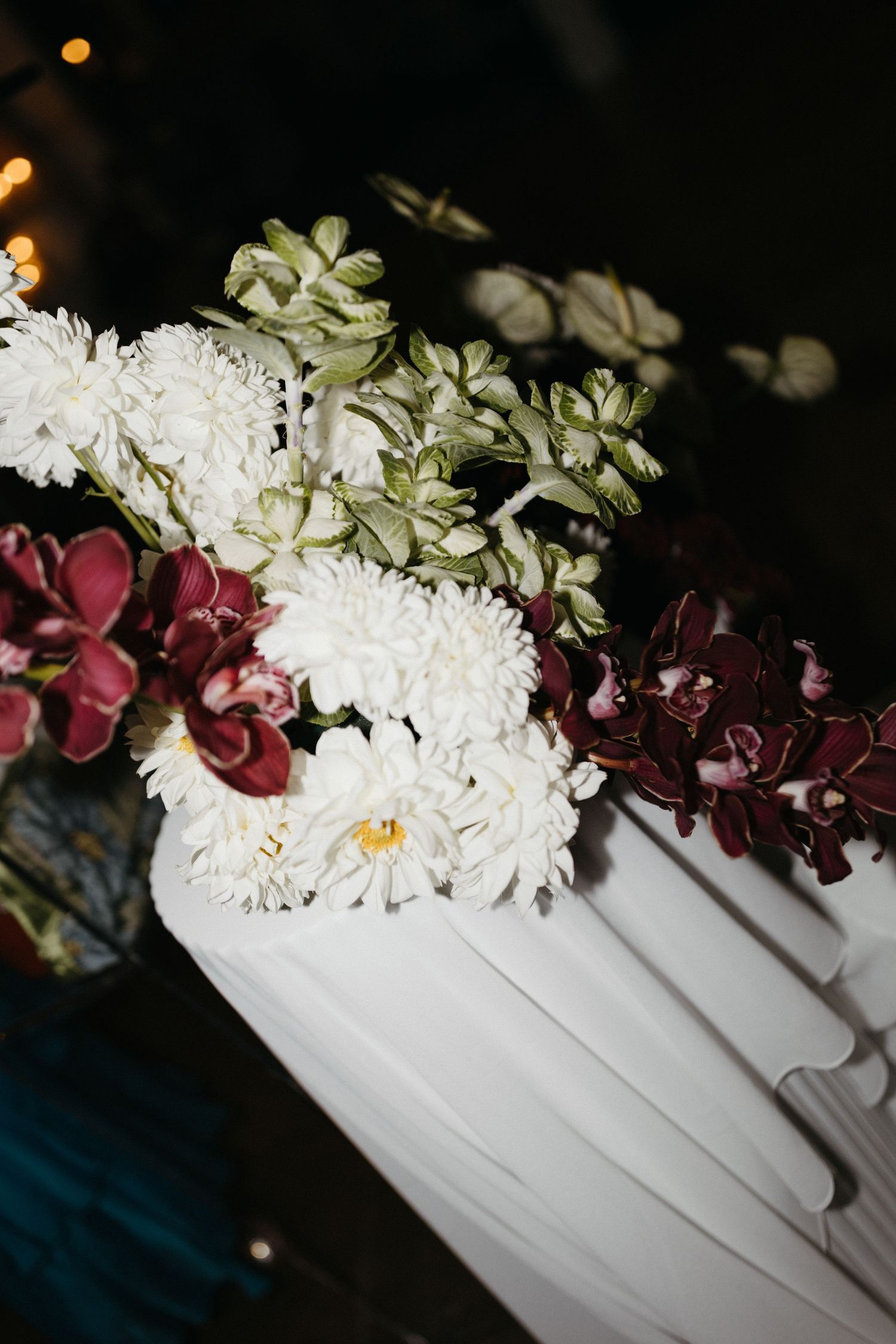 A floral arrangement featuring white chrysanthemums and burgundy orchids in a white vase against a dark background.