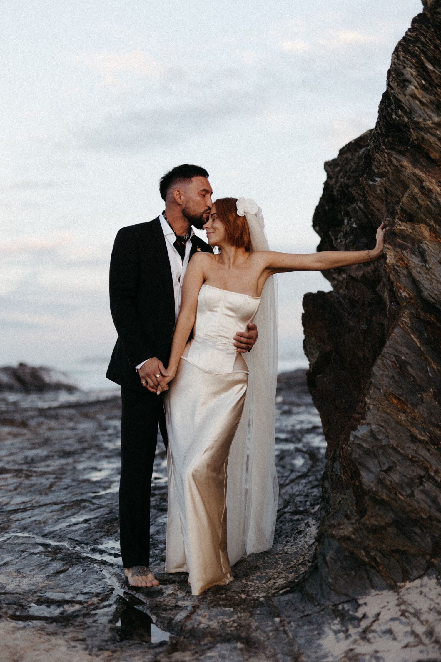 Couple in wedding attire embracing against dramatic coastal rock formation at sunset.
