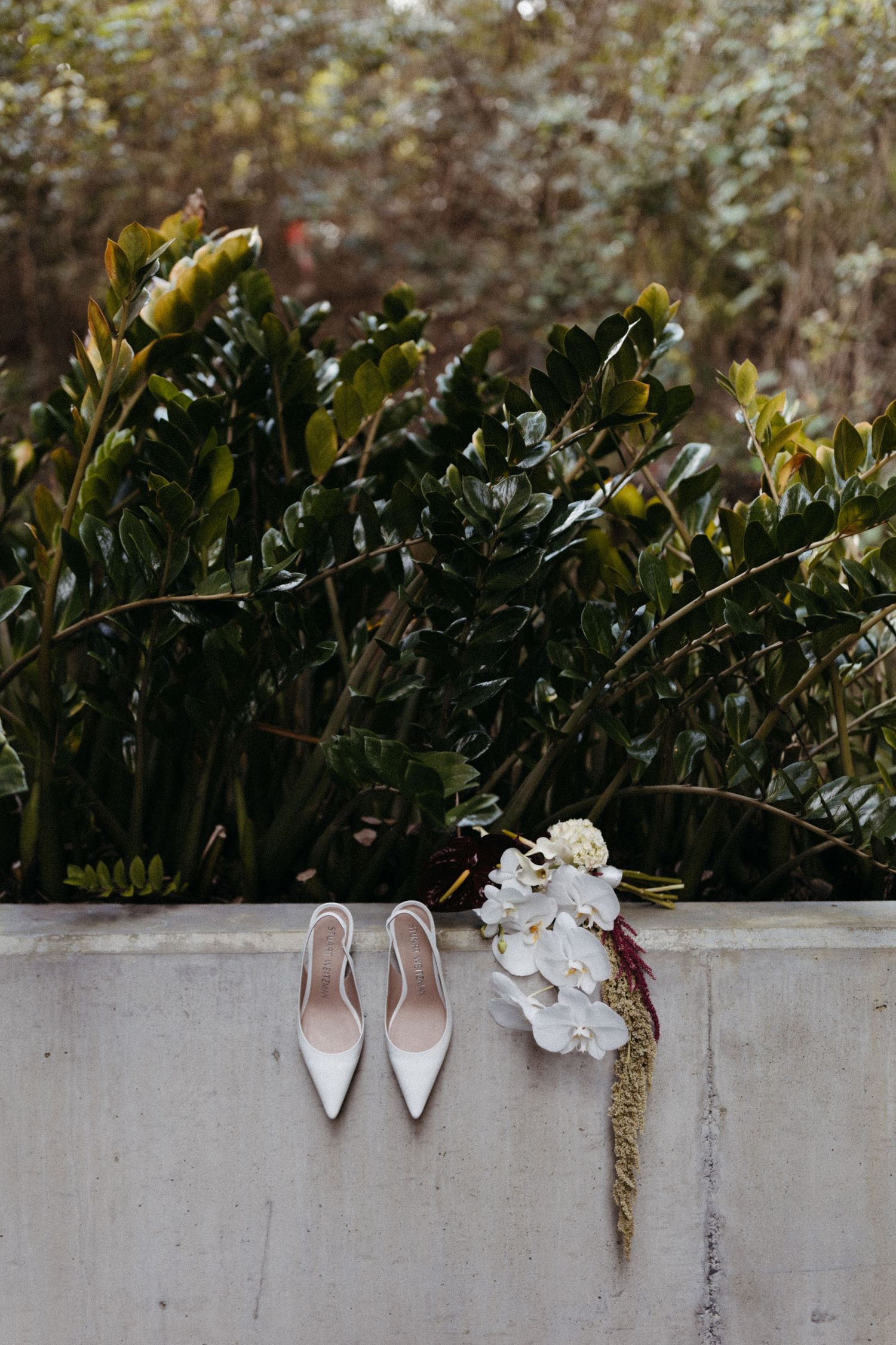 White bridal heels and a delicate floral bouquet arranged on a concrete ledge against lush green foliage backdrop.