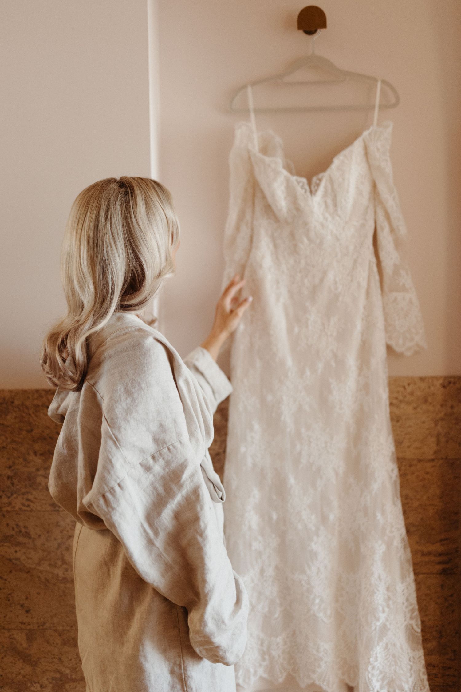 Someone wearing beige looks at an elegant white lace wedding dress with long sleeves hanging on display.