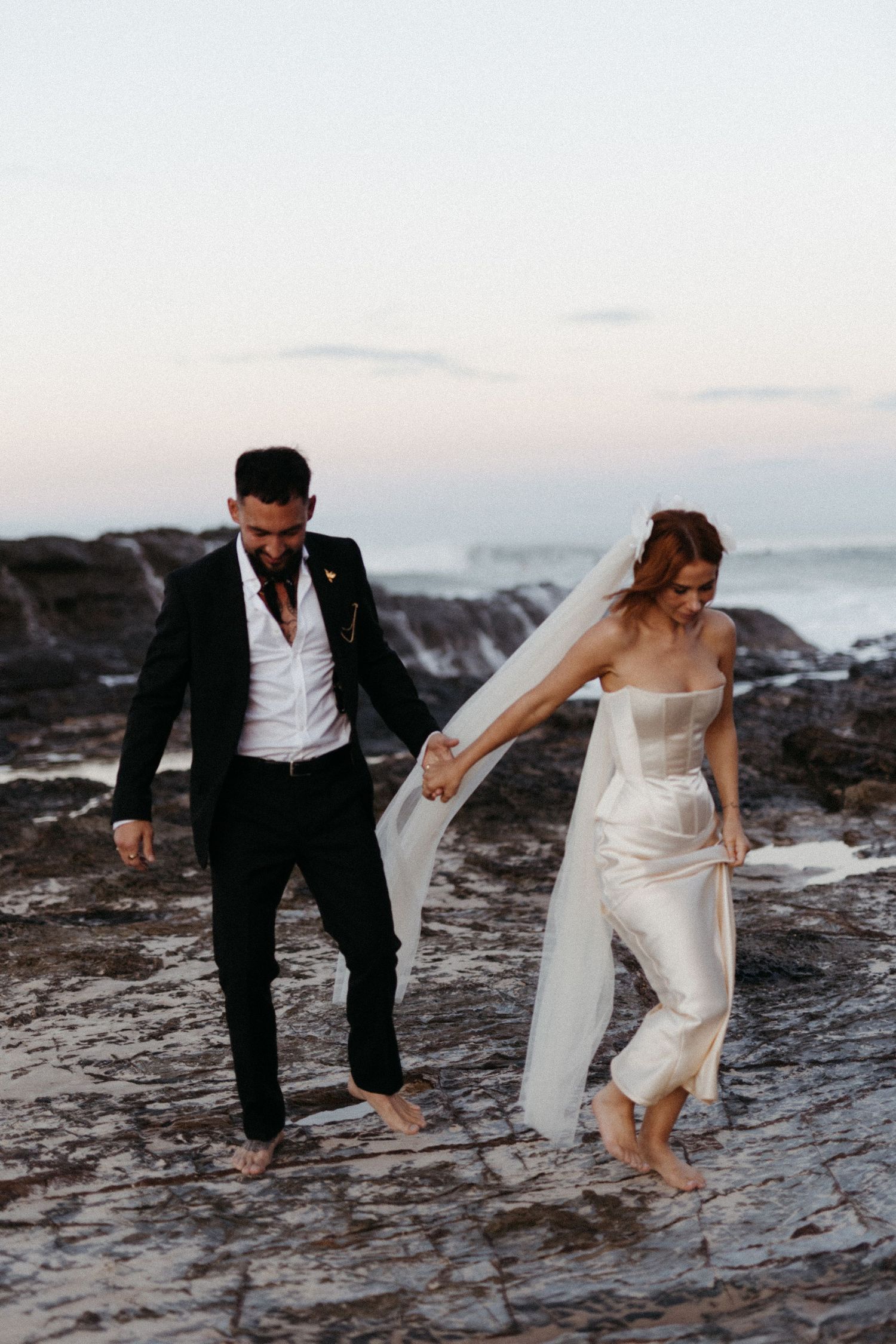 Newlywed couple exploring rocky beach at sunset as bride lifts white dress while walking over coastal terrain.