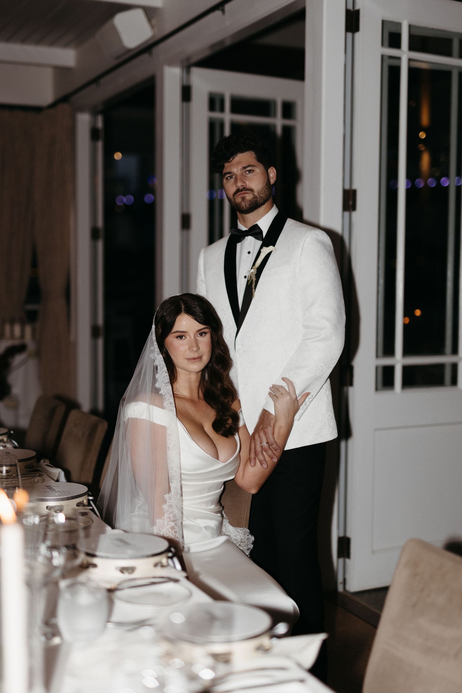 Wedding guests enjoy a candlelit evening reception at an elegant venue with white tablecloths and French doors.