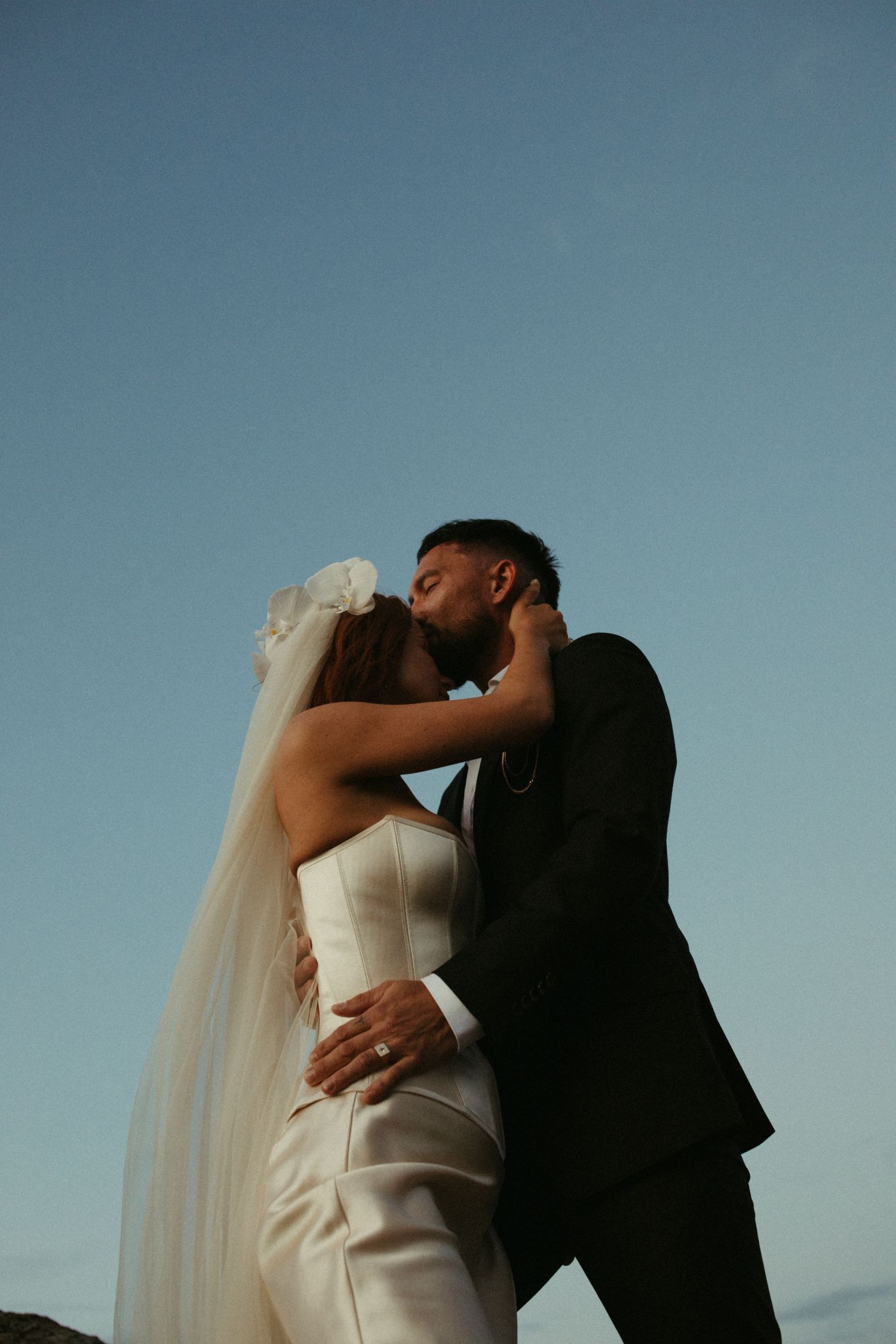Silhouetted newlyweds share intimate kisses against a blue sky in sequence of romantic wedding portraits.