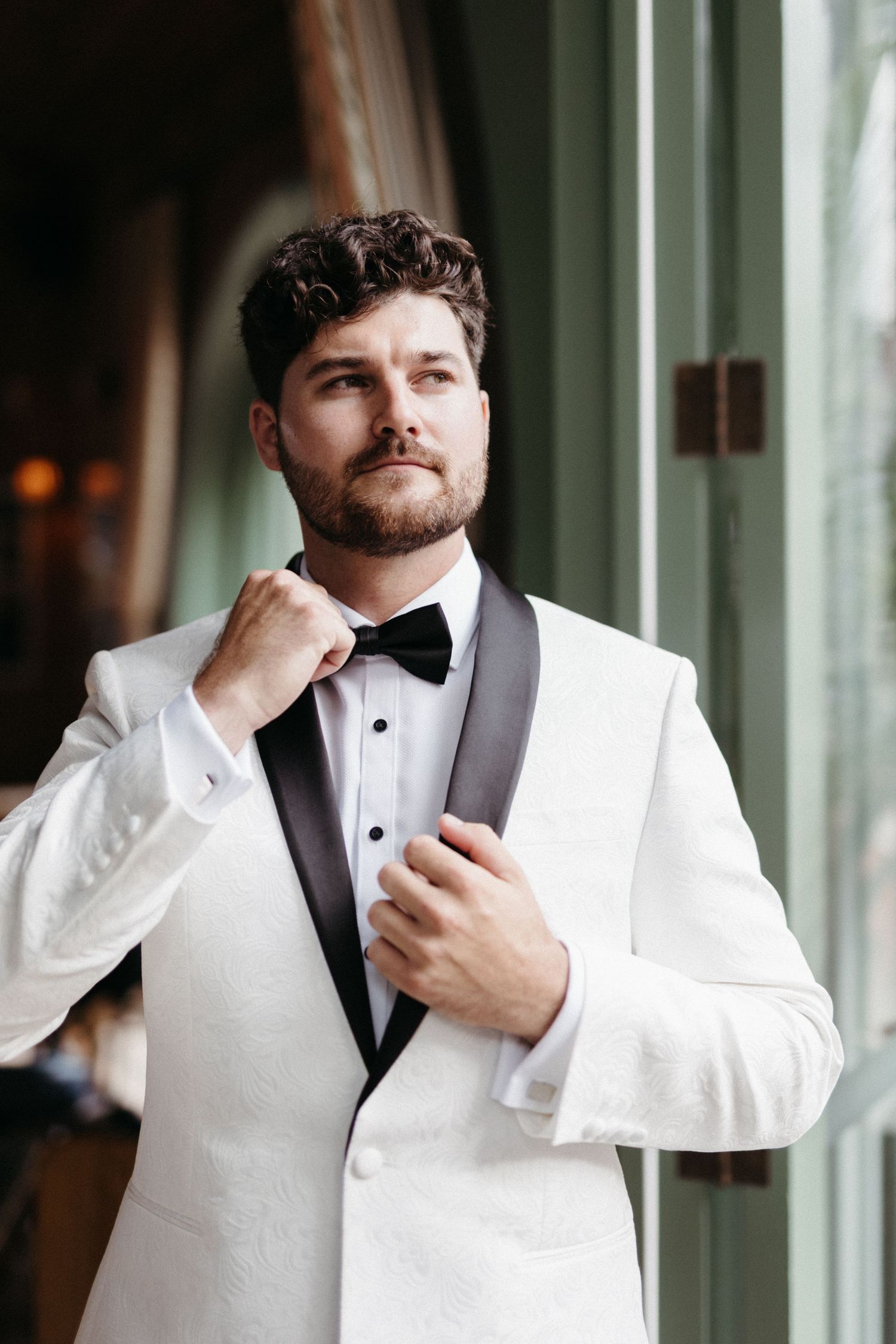 A person wearing a white tuxedo jacket with black lapels and bowtie adjusts their collar while standing by a window.