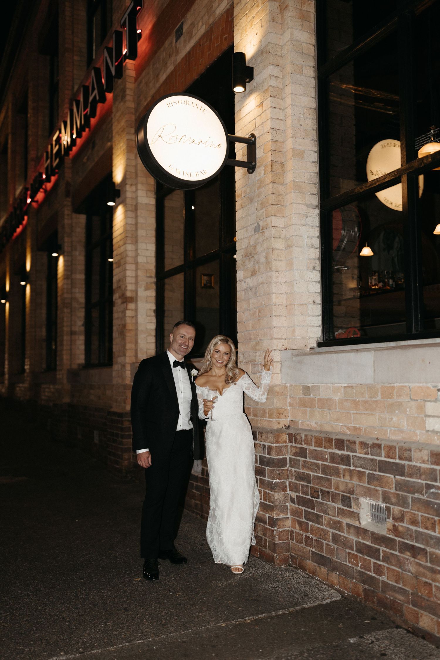 A formally dressed couple poses outside a restaurant with illuminated signage and brick walls at night.