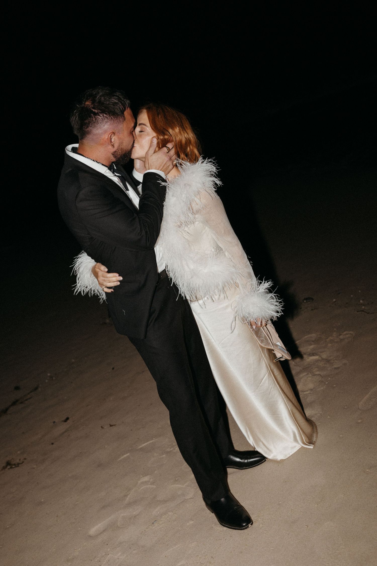 Romantic dip kiss between newlyweds on sandy beach at night.