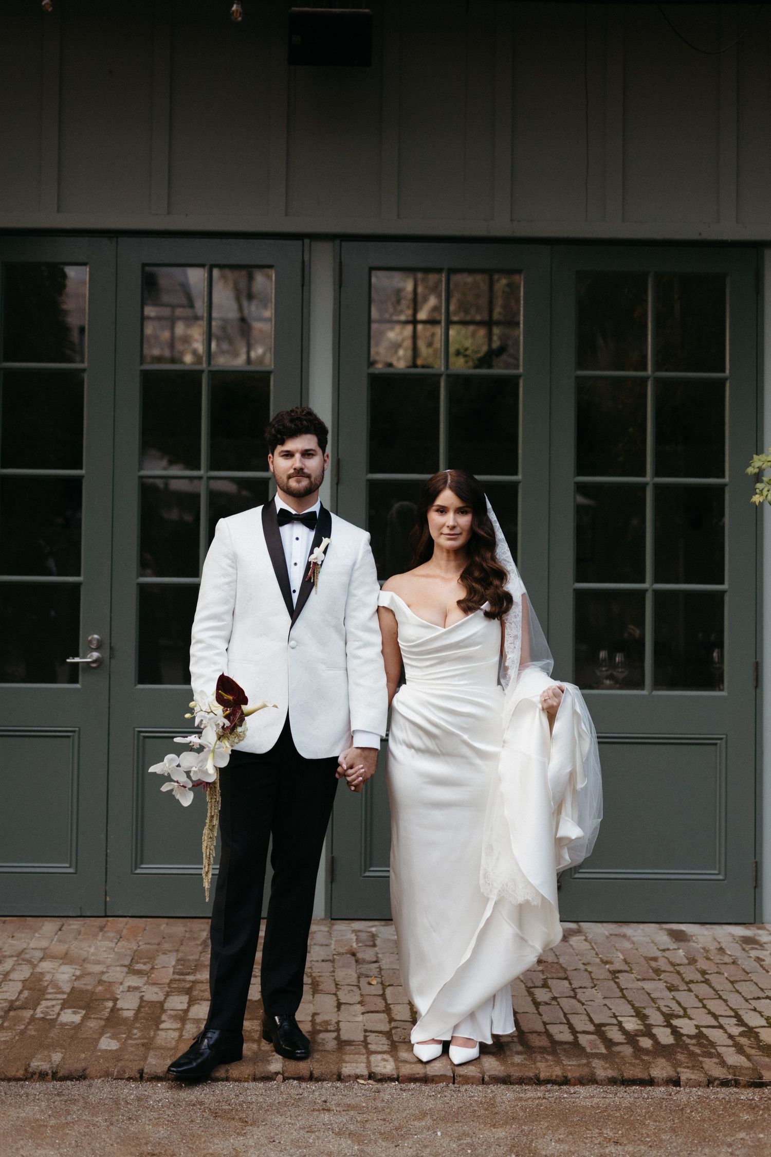A newlywed couple in formal wedding attire poses together against a gray building backdrop in a modern sequence.