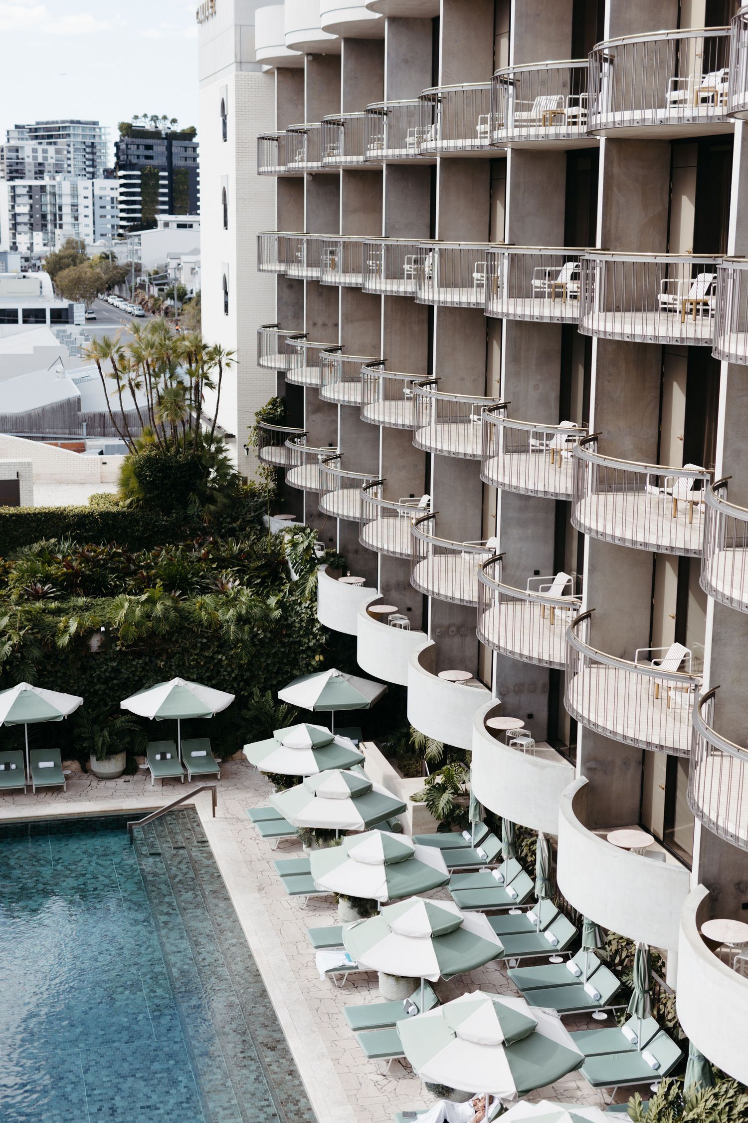 Modern hotel building with curved balconies overlooks a pool area lined with turquoise lounge chairs and white umbrellas.