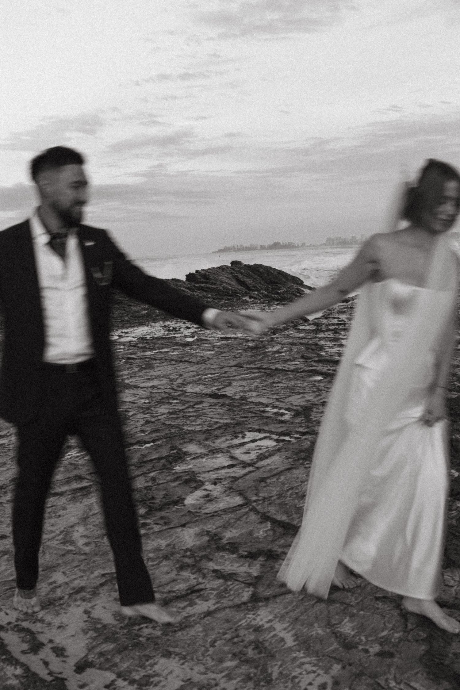 Wedding couple dances joyfully on rocky terrain with dramatic sky and cityscape in background.