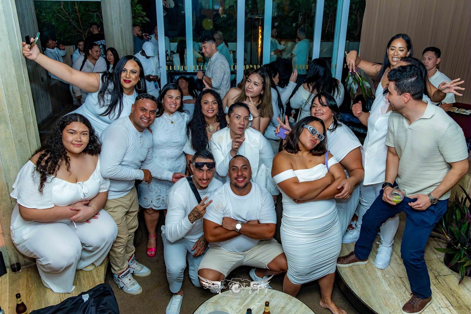 Large group wearing all white outfits gathered together for celebratory photo indoors.