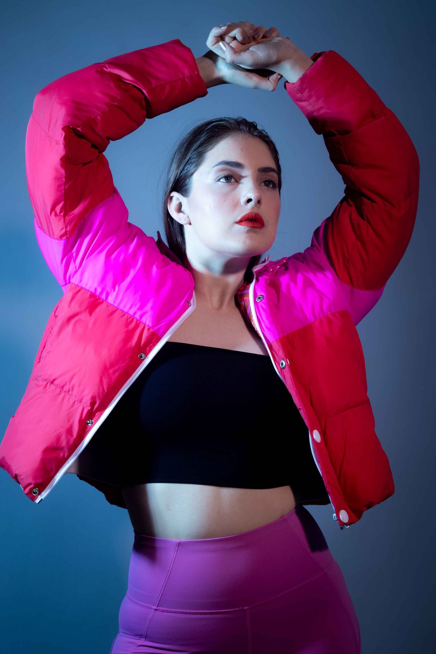 Fashion portrait featuring red puffer jacket and purple shorts against blue backdrop.