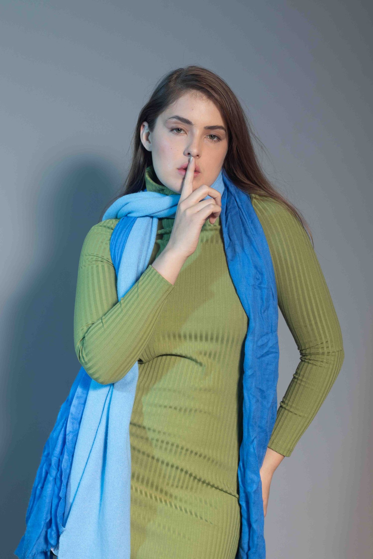 Portrait in olive green ribbed dress with blue scarf making hushing gesture.