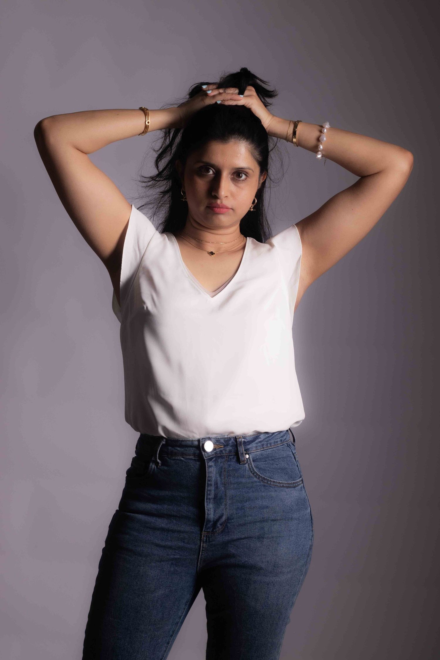 Portrait of person in casual white top and jeans fixing hair against gray background.
