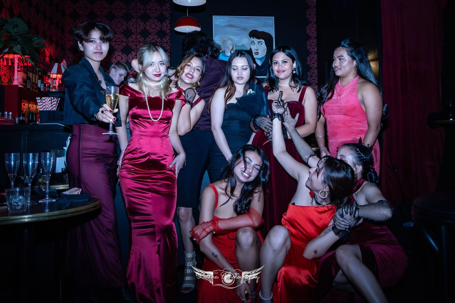Group poses in glamorous evening attire at a nightclub with patterned wallpaper and mood lighting.