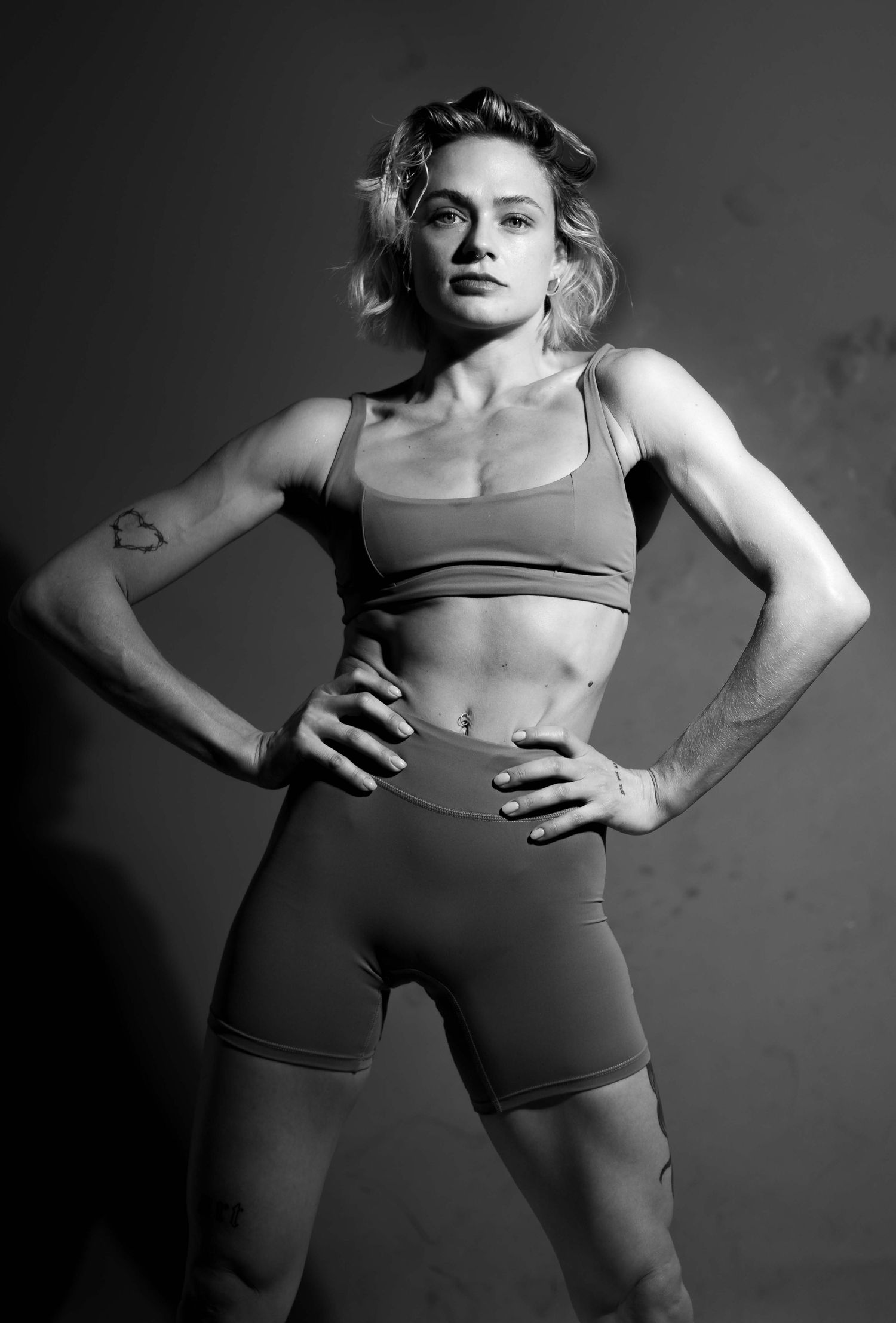 Athletic figure in black and white wearing sports bra and shorts poses with hands on hips against dark background.