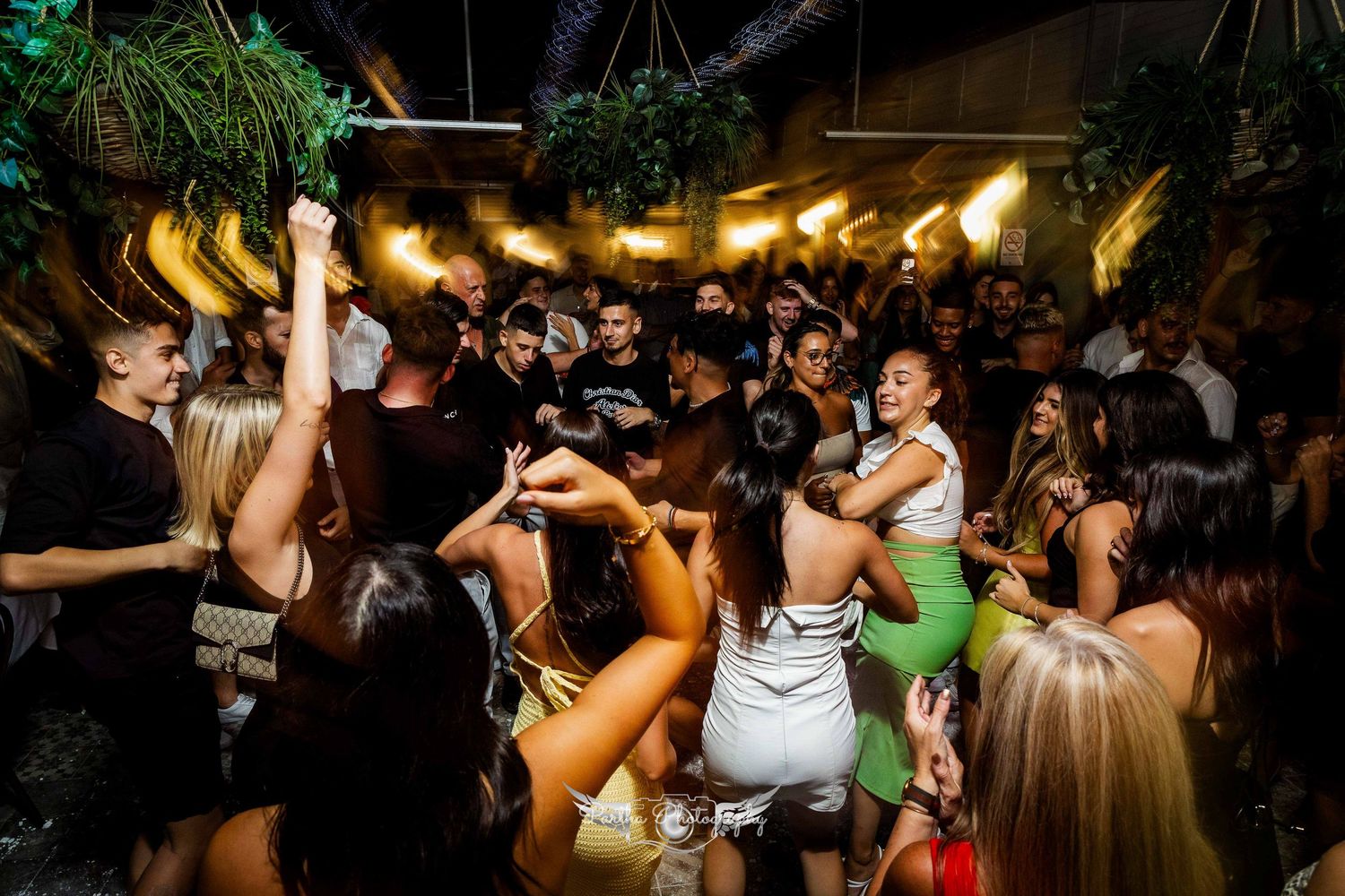 Crowded nightclub dance floor with partygoers dancing under tropical decor and warm lighting.