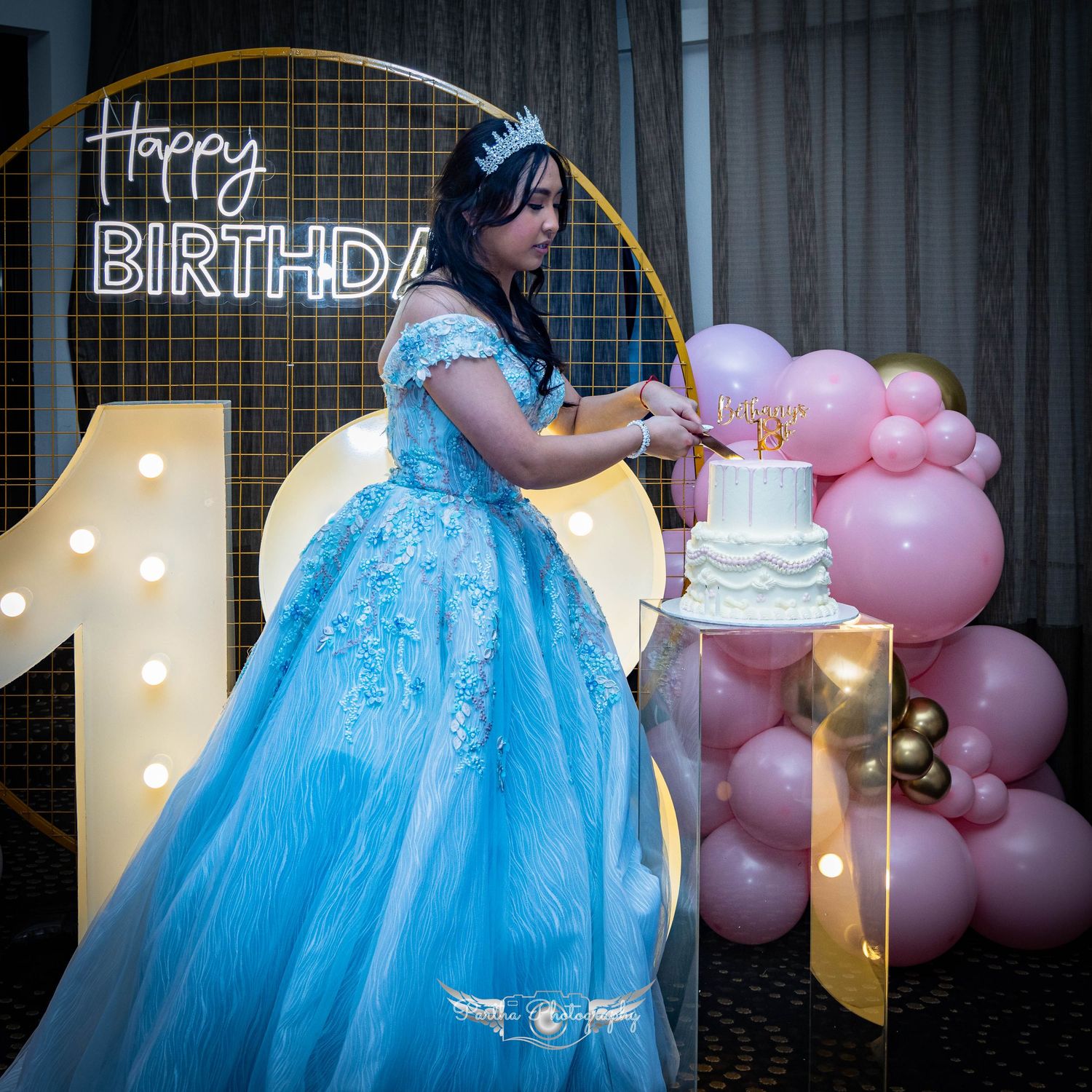 A princess in a blue ballgown poses next to a decorated birthday cake and pink balloons.