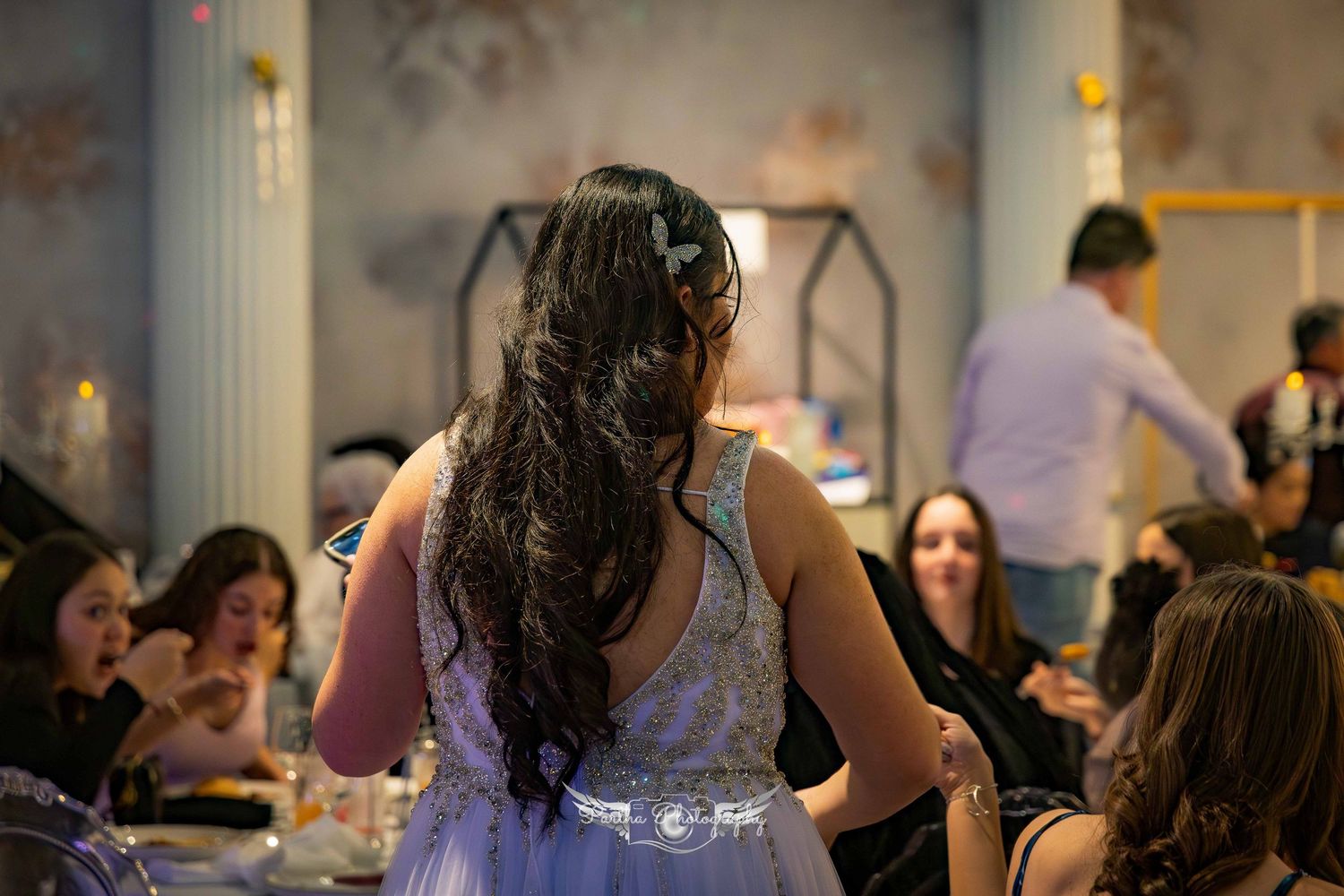 A woman with long dark hair in a formal dress stands among seated guests at an event.