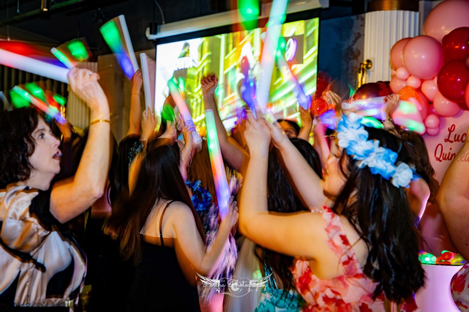 Party guests dancing and holding colorful light sticks during a celebration with balloon decorations.