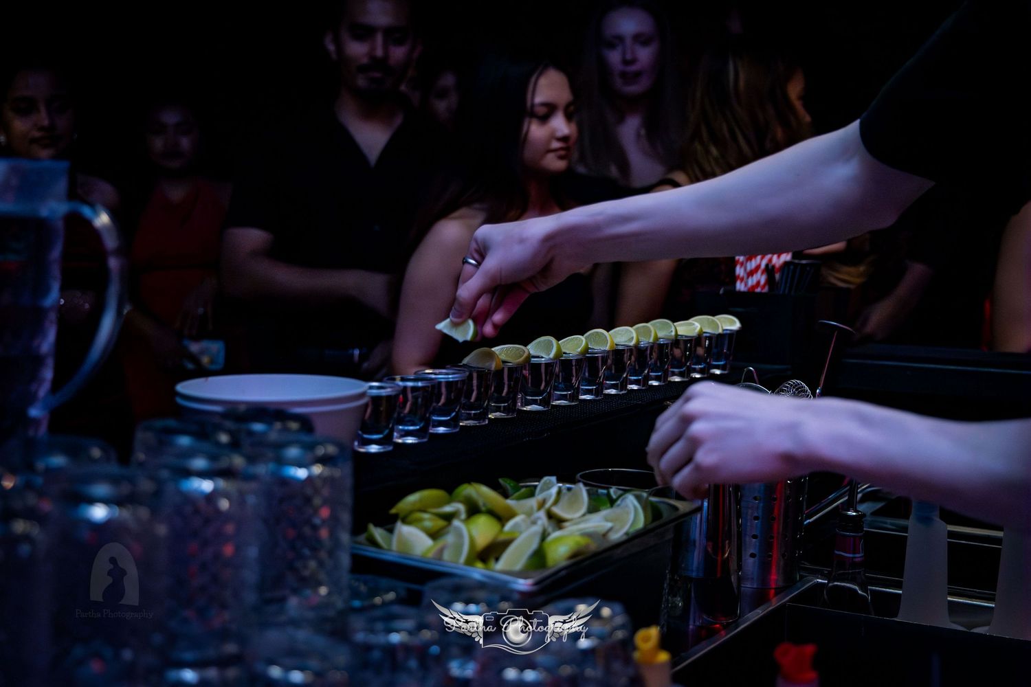 Close-up of hands pouring drinks at a bar with lemon garnishes and glassware in dim lighting.