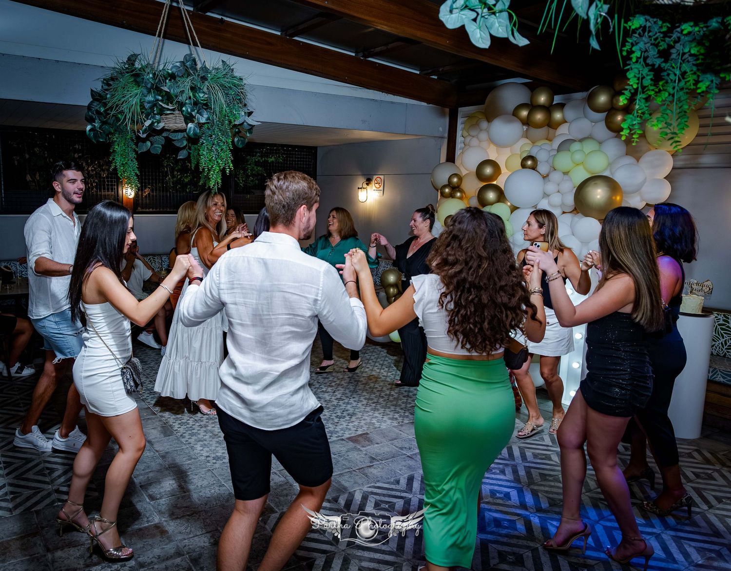 People dancing on geometric patterned floor with green plants and balloons overhead.