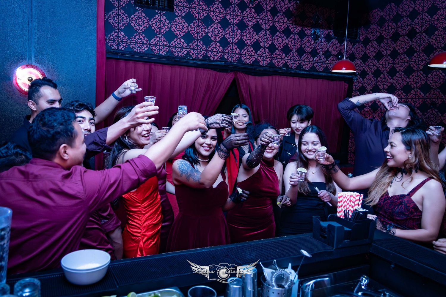 A crowded bar scene with people reaching across the counter holding drinks in red and dark lighting.