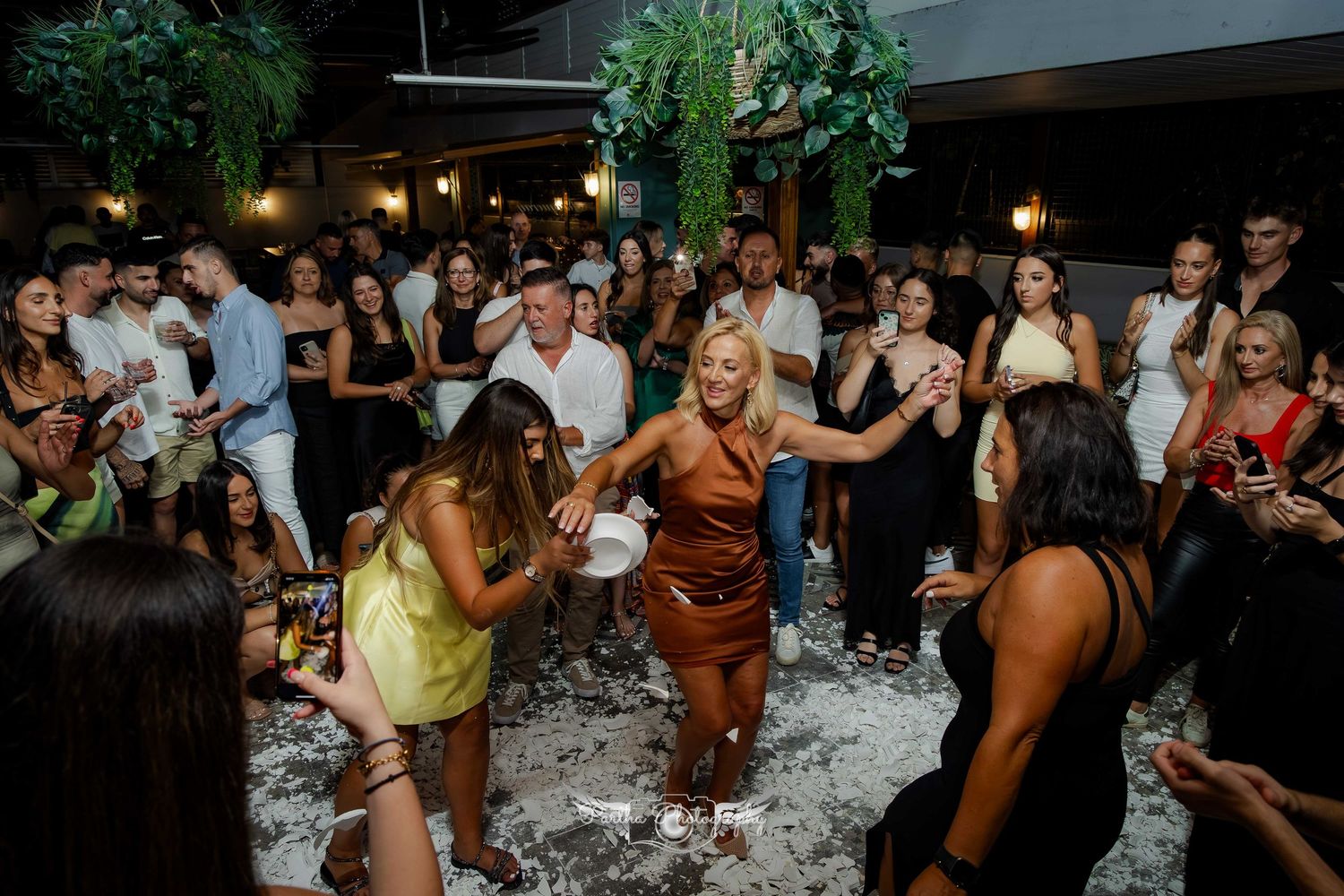Crowd of partygoers dancing and celebrating in a venue decorated with hanging greenery.
