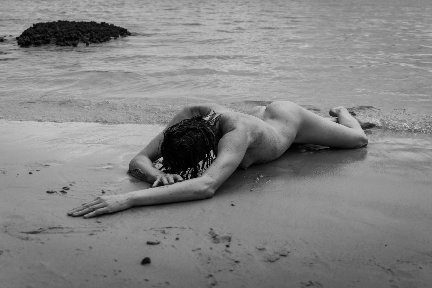 Black and white artistic nude reclining on beach with waves in background.