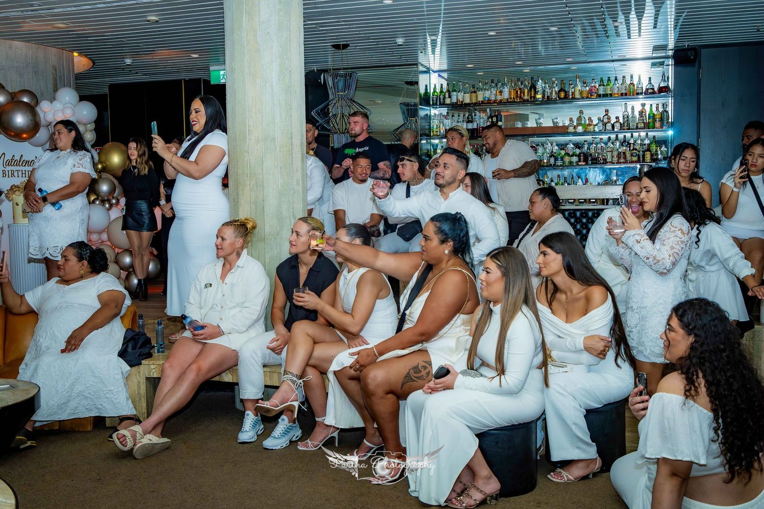 Party guests in white attire dancing and celebrating in venue with bar area.