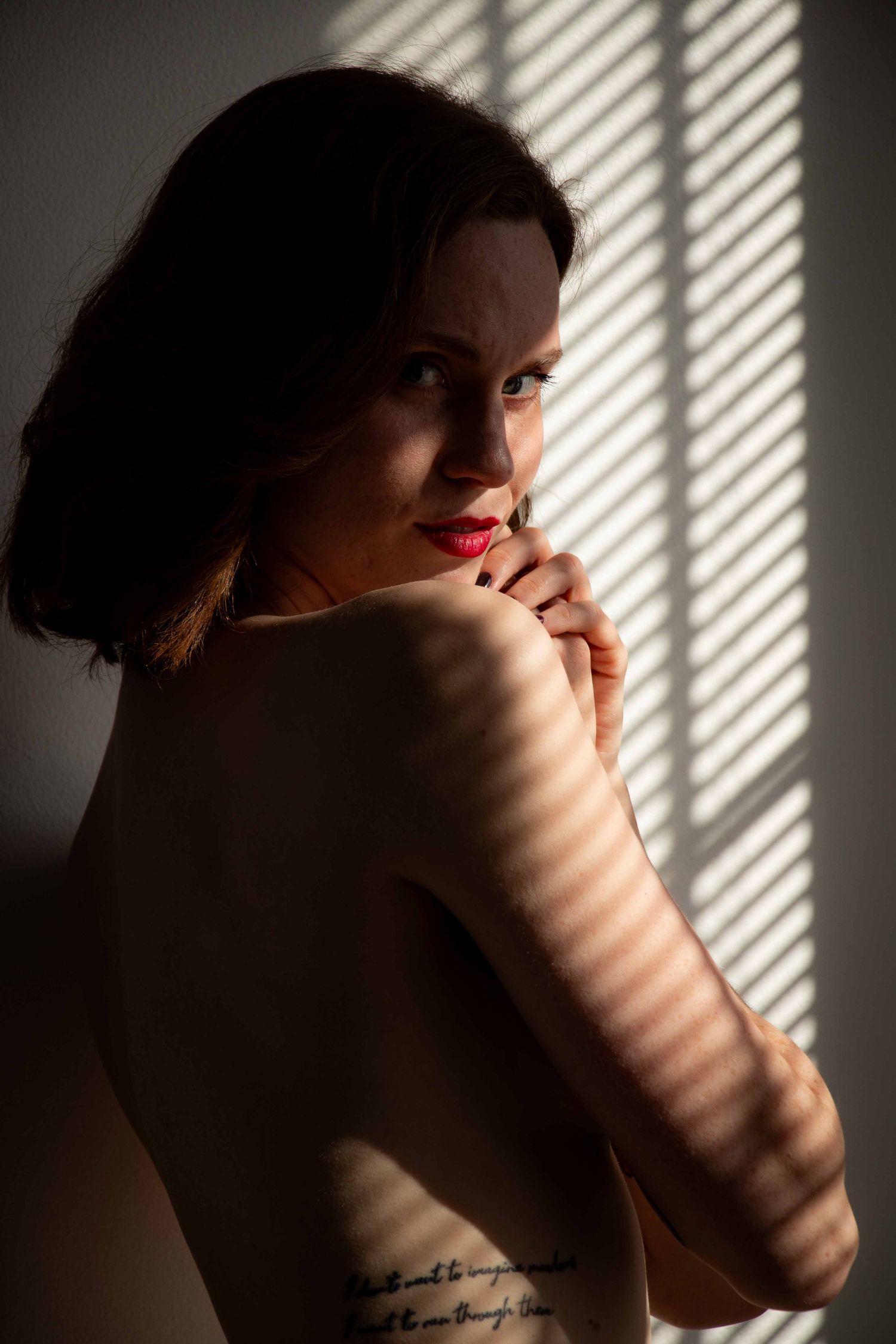 Dramatic portrait with venetian blind shadows creating stripes across bare shoulder.