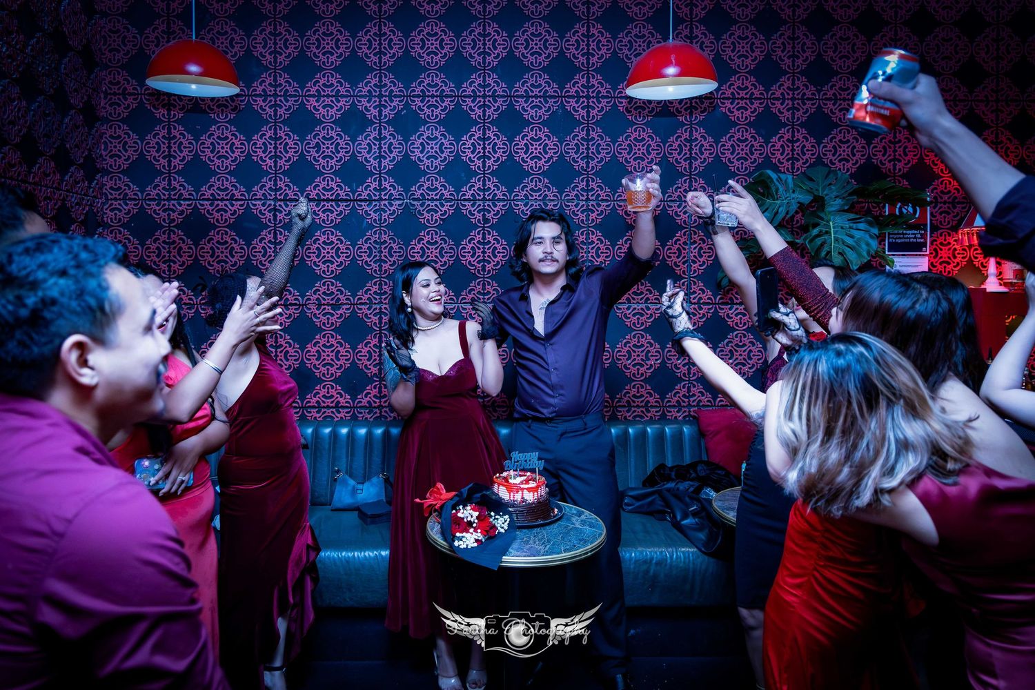 Group of revelers dancing and celebrating in a nightclub with red pendant lights and patterned wallpaper.